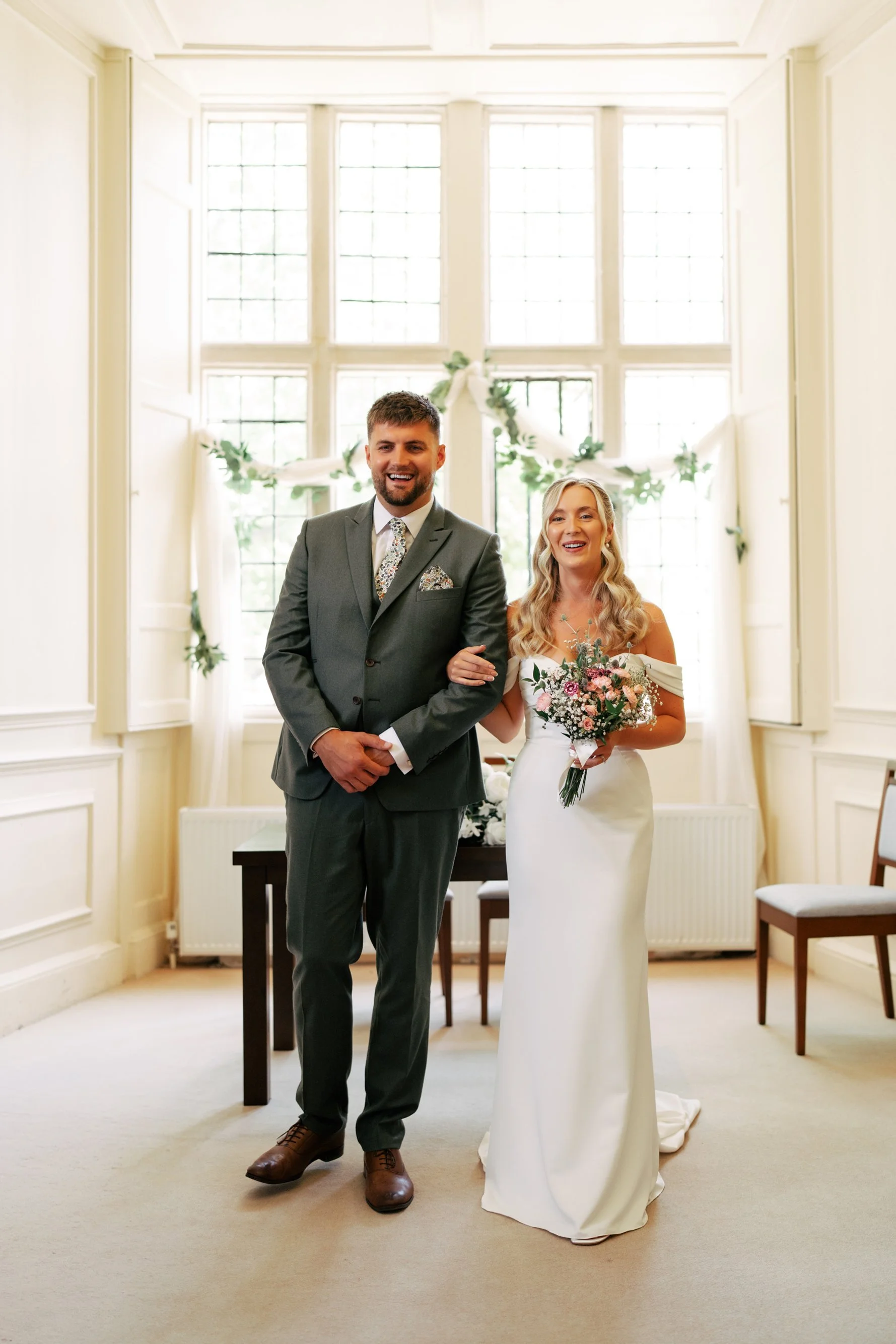 A couple dressed in wedding attire standing inside a bright room with large windows and white walls. The man wears a gray suit with a floral tie, and the woman wears a white off-shoulder wedding dress holding a bouquet of flowers.