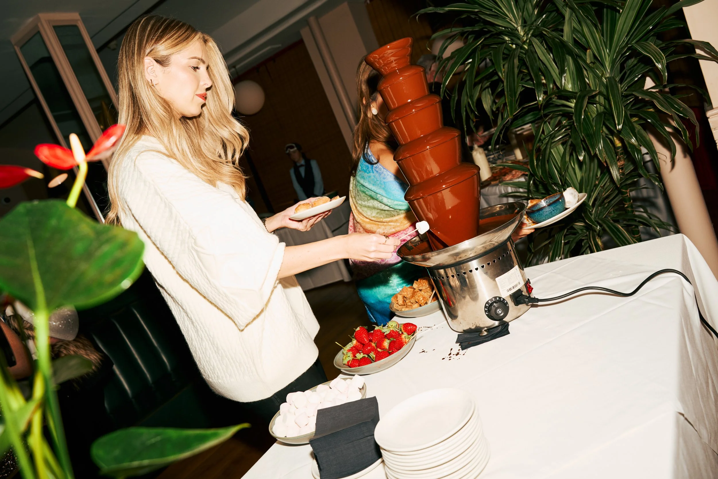 A woman in a white sweater serving herself chocolate fondue from a stack of bowls at a buffet table with strawberries, marshmallows, and other treats.
