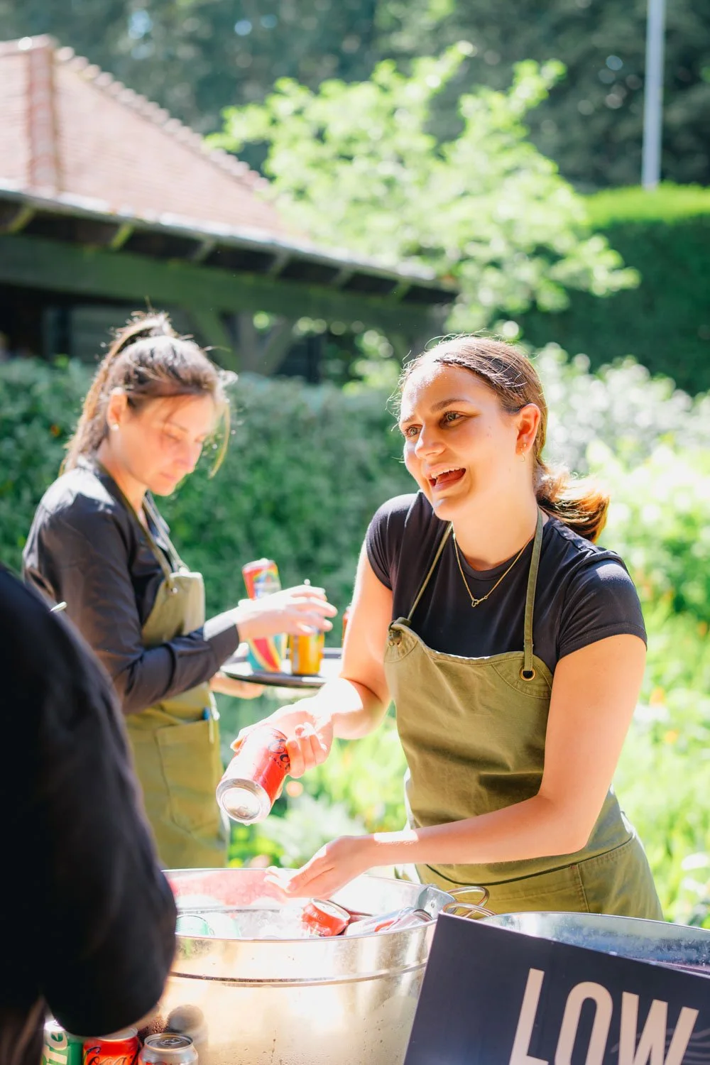 Two women outdoors at a summer gathering, one serving drinks from cans into a large bowl, both wearing aprons, with greenery and a house in the background.