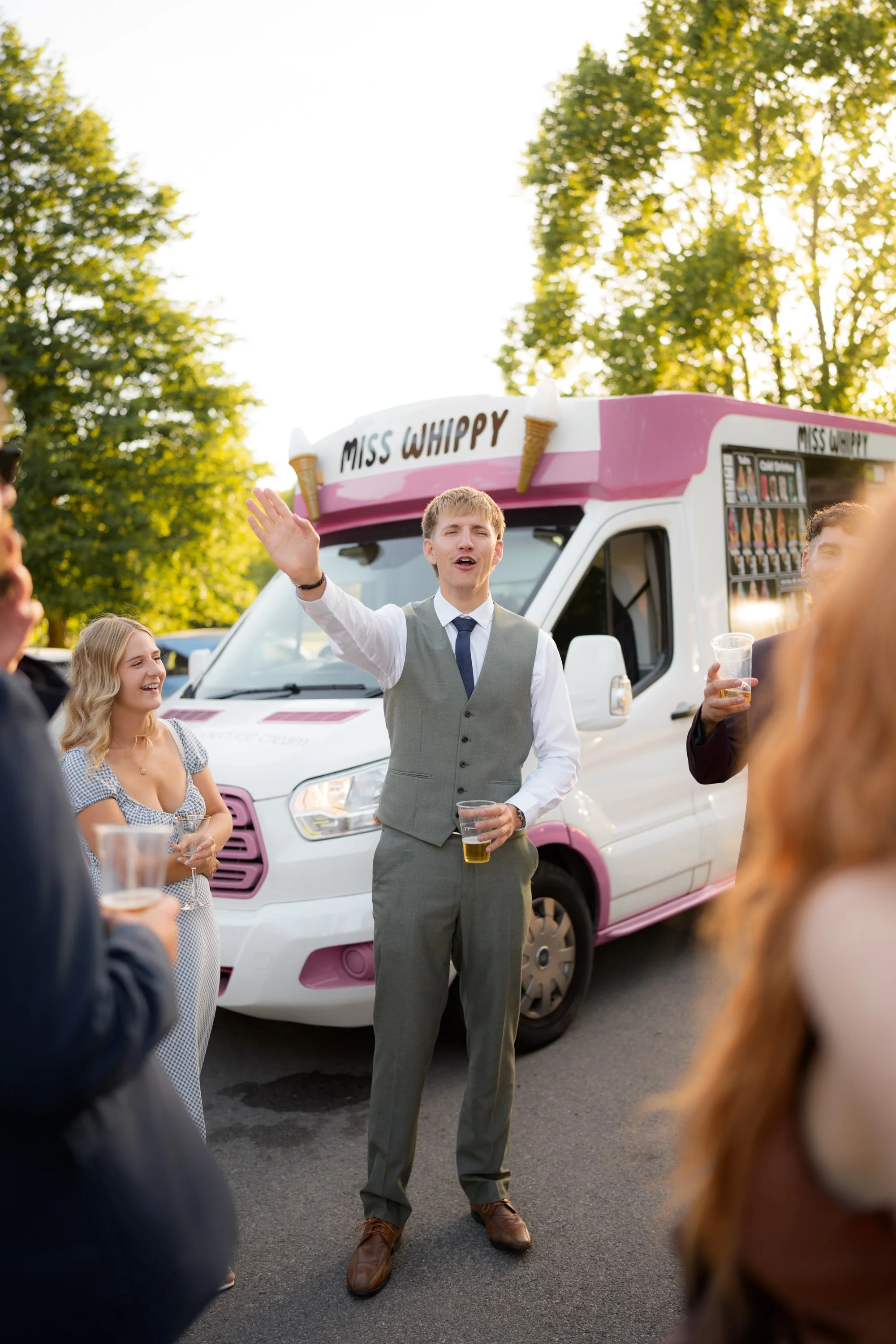 A man in a gray vest and tie raising his hand while holding a drink stands in front of a pink and white ice cream truck labeled 'Miss Whippy' at an outdoor event. Several people are around him, some holding drinks, with trees and sunlight in the back