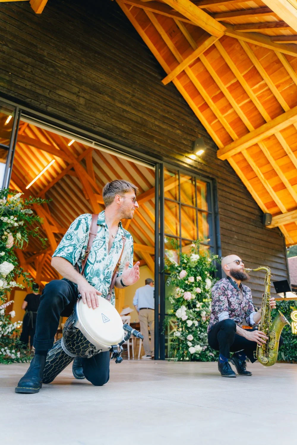 Two musicians performing in front of a rustic wooden building with an open sliding glass door, decorated with flowers and greenery. One musician is playing a hand drum, and the other is playing a saxophone.