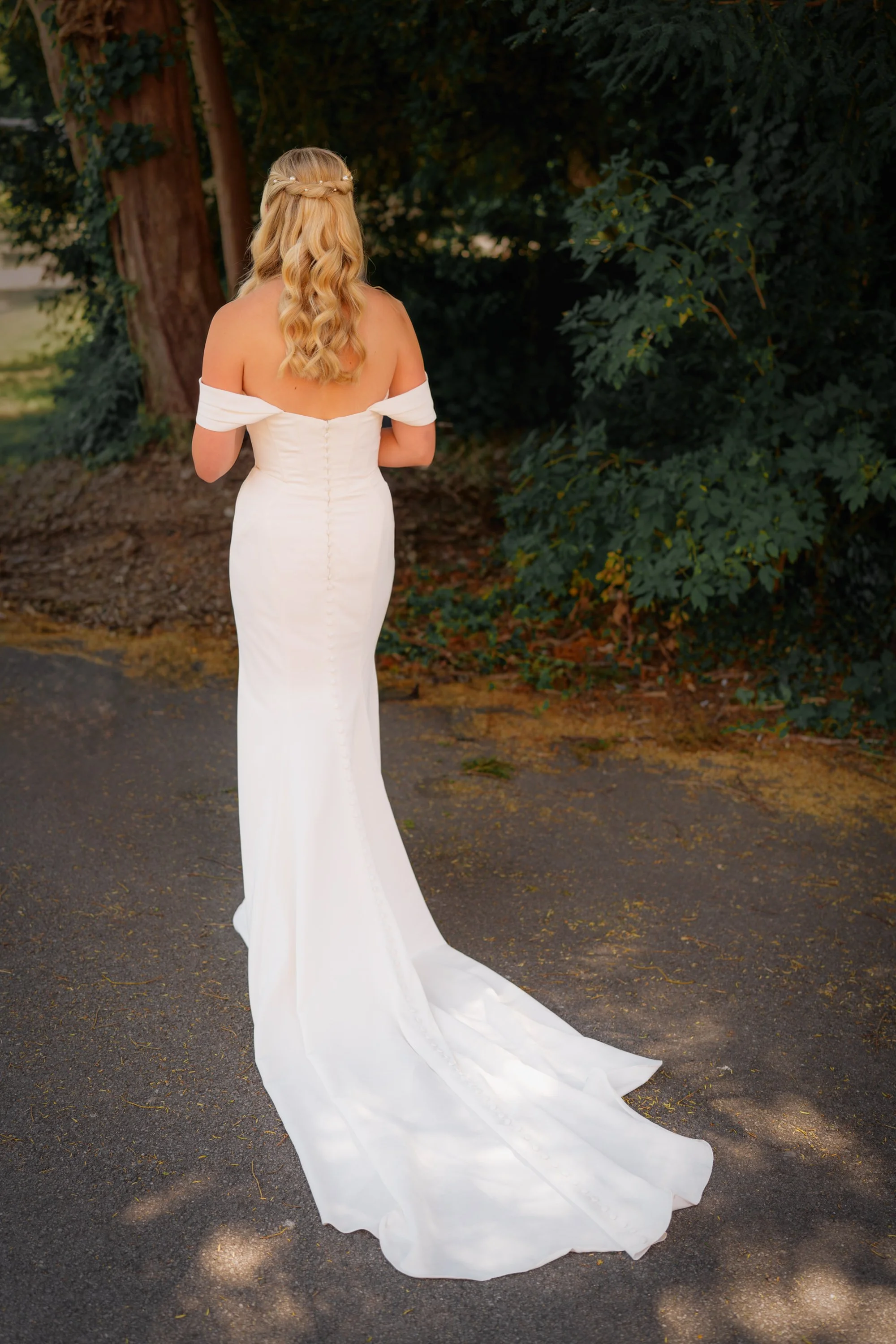 A woman with long blonde curly hair wearing an off-shoulder white wedding dress with a long train, standing outdoors in front of large dark green trees.