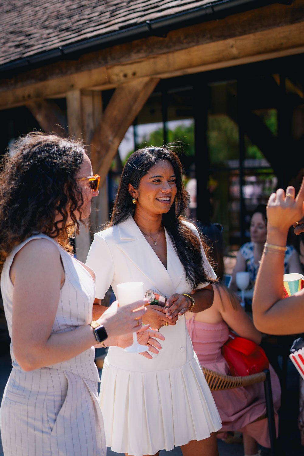 Two women are talking at an outdoor gathering, one holding a drink, both smiling and dressed in stylish summer outfits, with other guests in the background.
