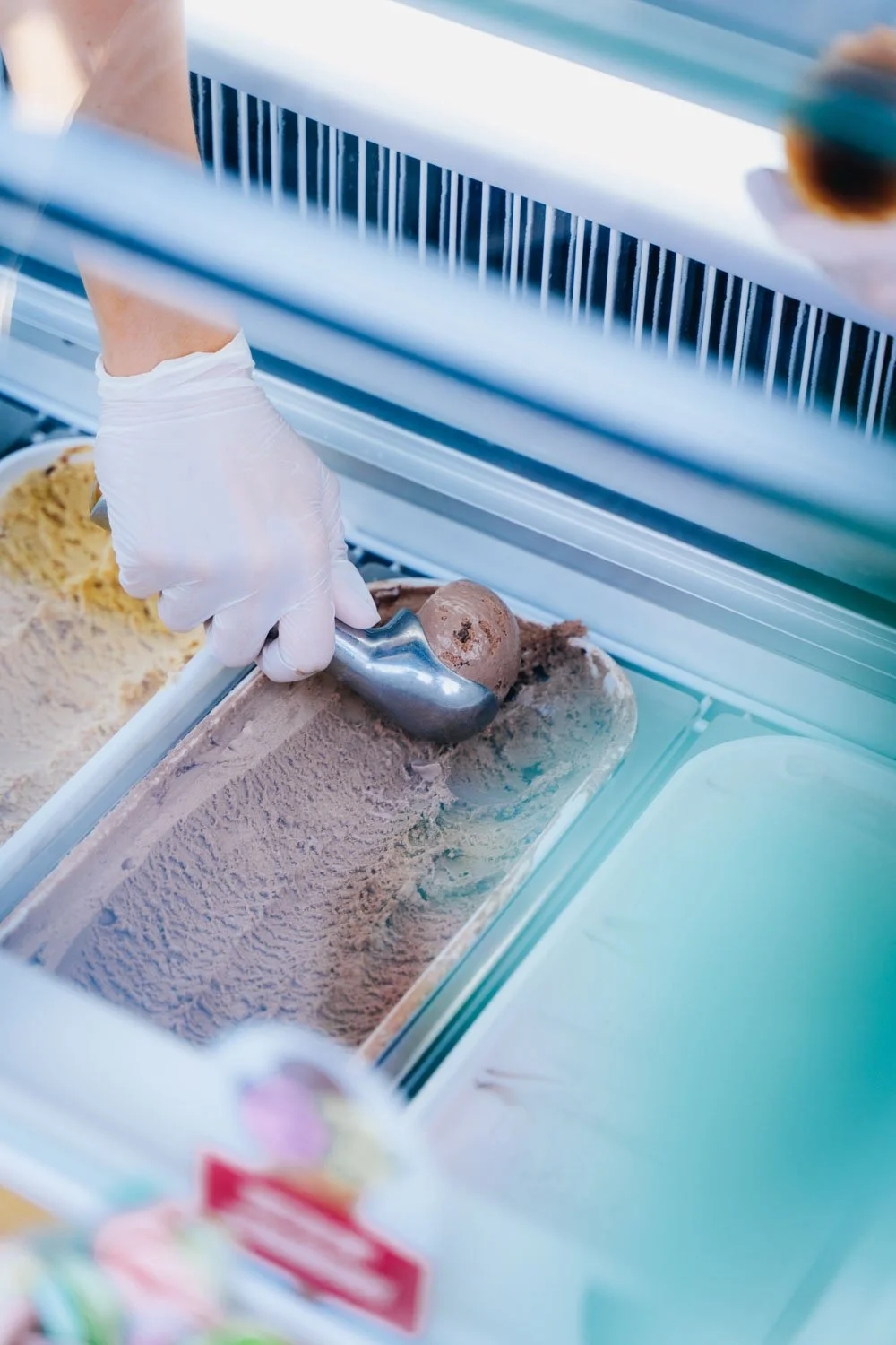 A person wearing a white glove scoops chocolate ice cream from a metal container in an ice cream shop freezer.