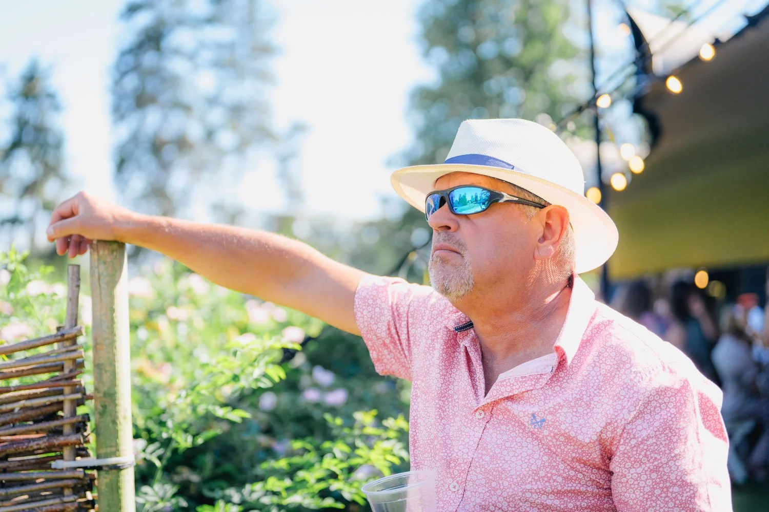 A man wearing a white hat and sunglasses standing outdoors, reaching for an object, with a blurred background of people and trees.