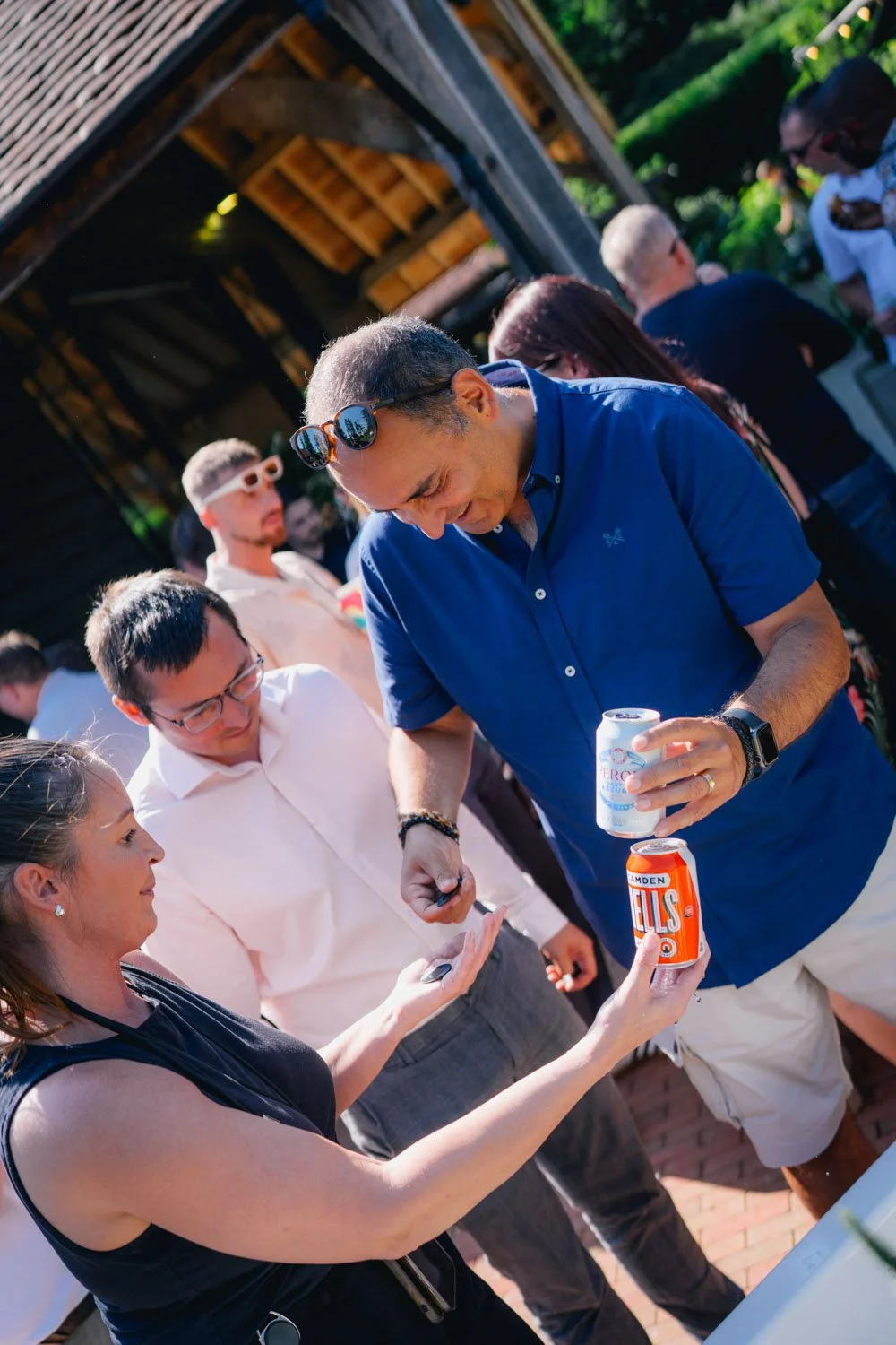 People at an outdoor gathering, chatting and holding drinks, under a wooden pavilion with string lights.