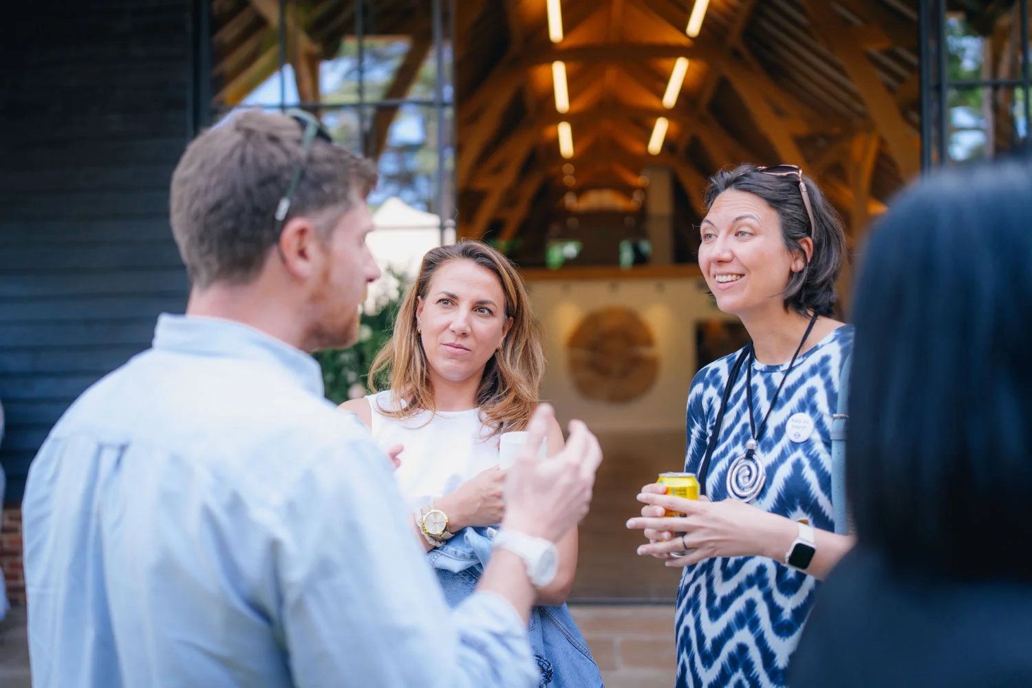 Four people engaged in conversation at a gathering outside a building with a wooden interior and modern lighting. The group includes two women and two men, with one woman smiling and holding a beverage, and the others listening and gesturing.