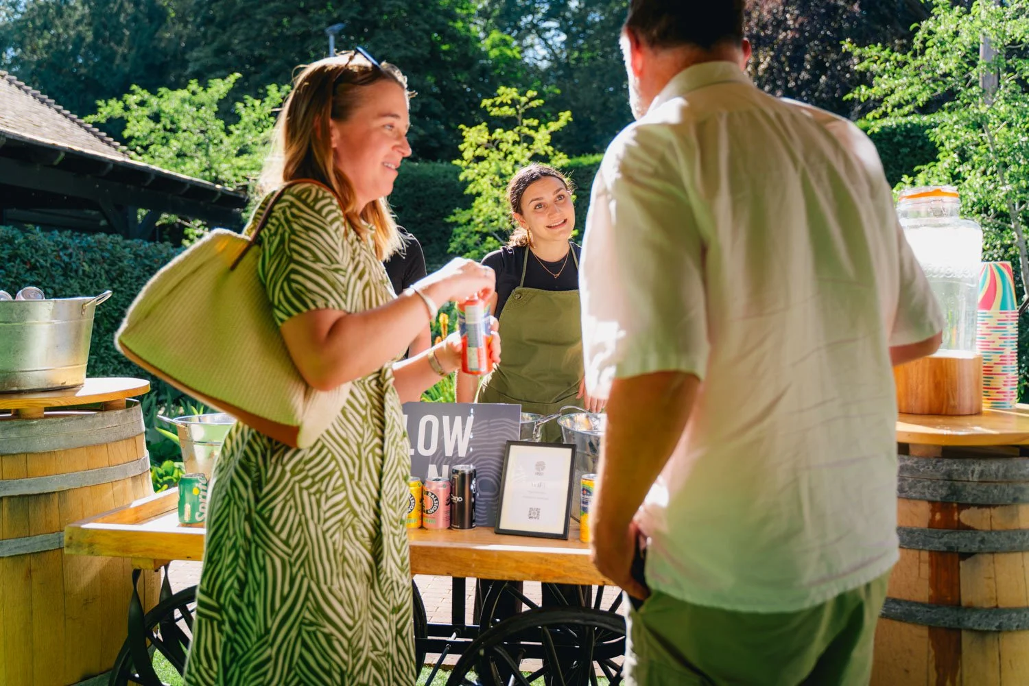 Three people engaging in conversation at an outdoor event with a table displaying drinks and promotional materials, bright sunlight, trees, and a building in the background.