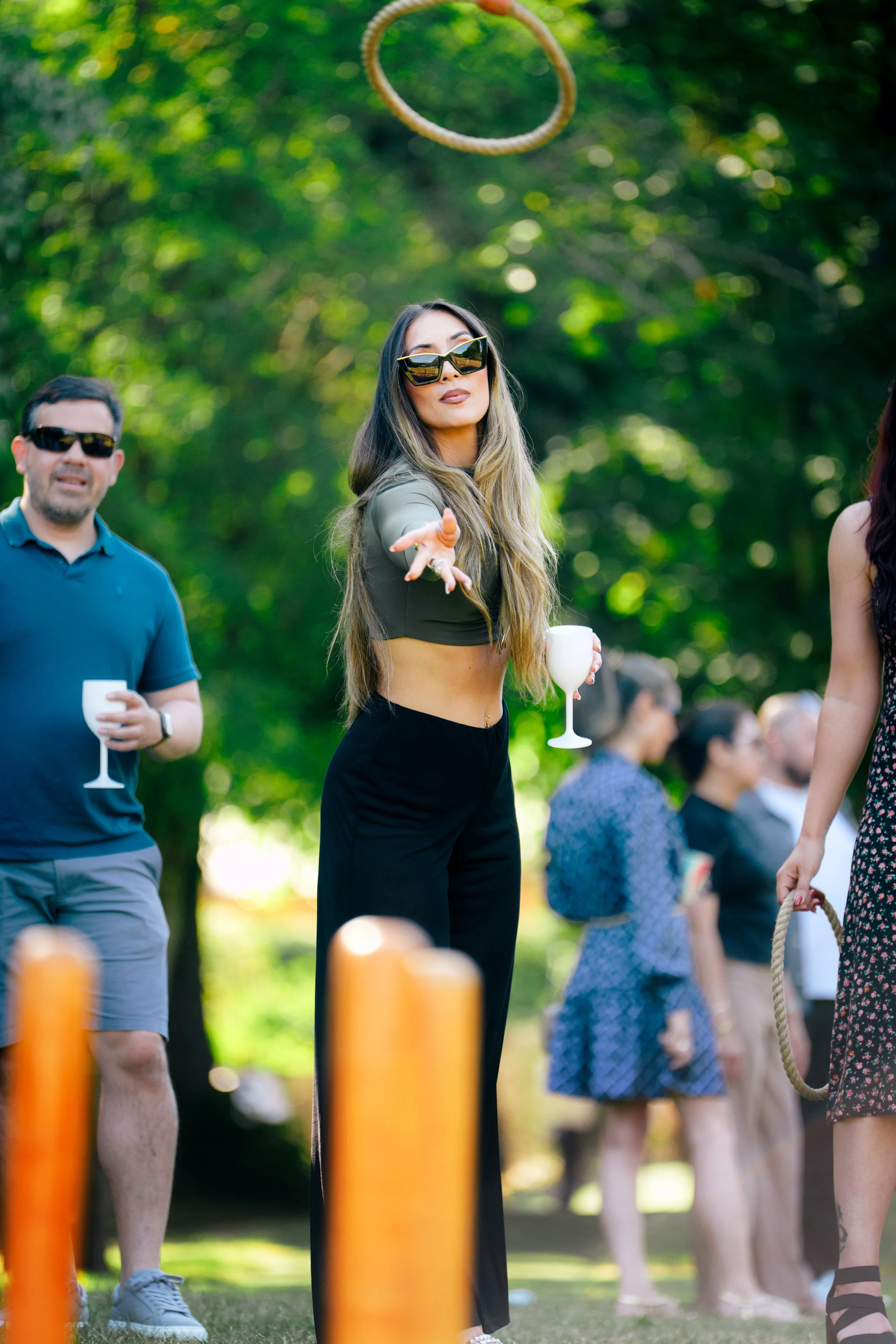 Group of people outdoors, with a woman in the center reaching out, wearing sunglasses and holding a wine glass, with others in background, some holding drinks, during daytime.