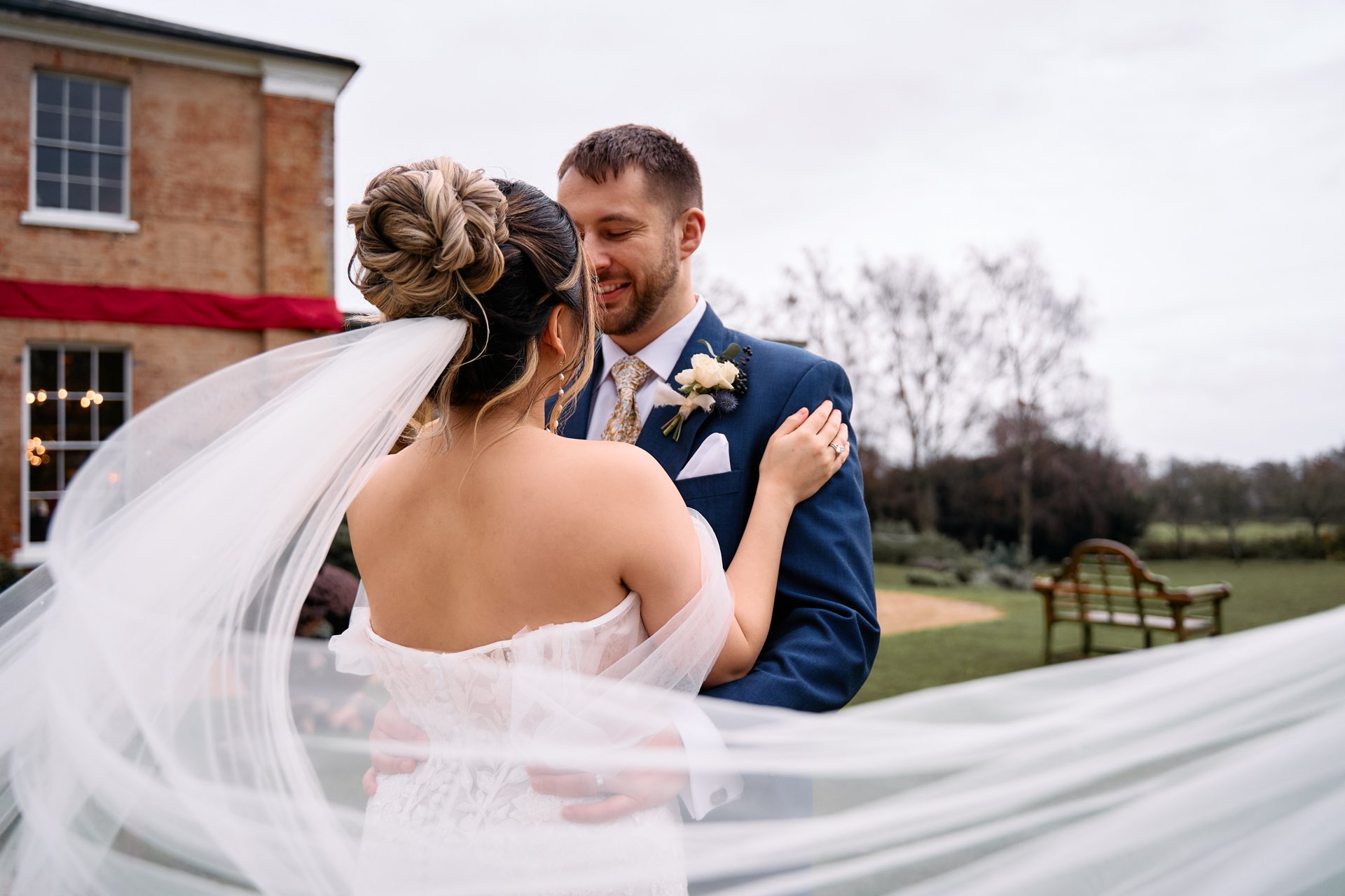 A bride and groom share a moment outdoors on their wedding day, with the bride in a white dress and veil and the groom in a navy suit with a boutonnière, smiling at each other.