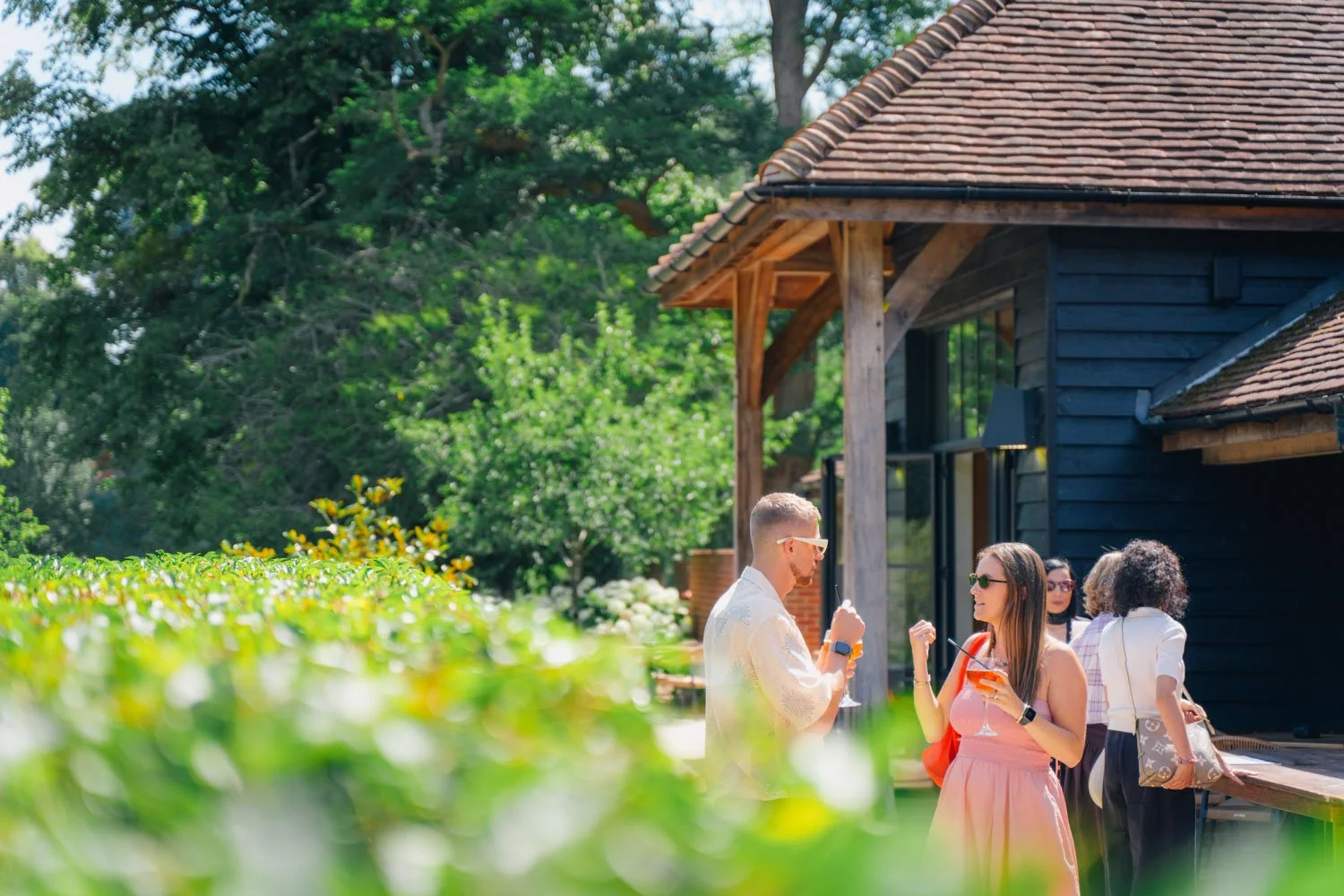 Group of four people enjoying drinks and conversation outdoors near a black wooden house with a tiled roof, surrounded by greenery and trees.