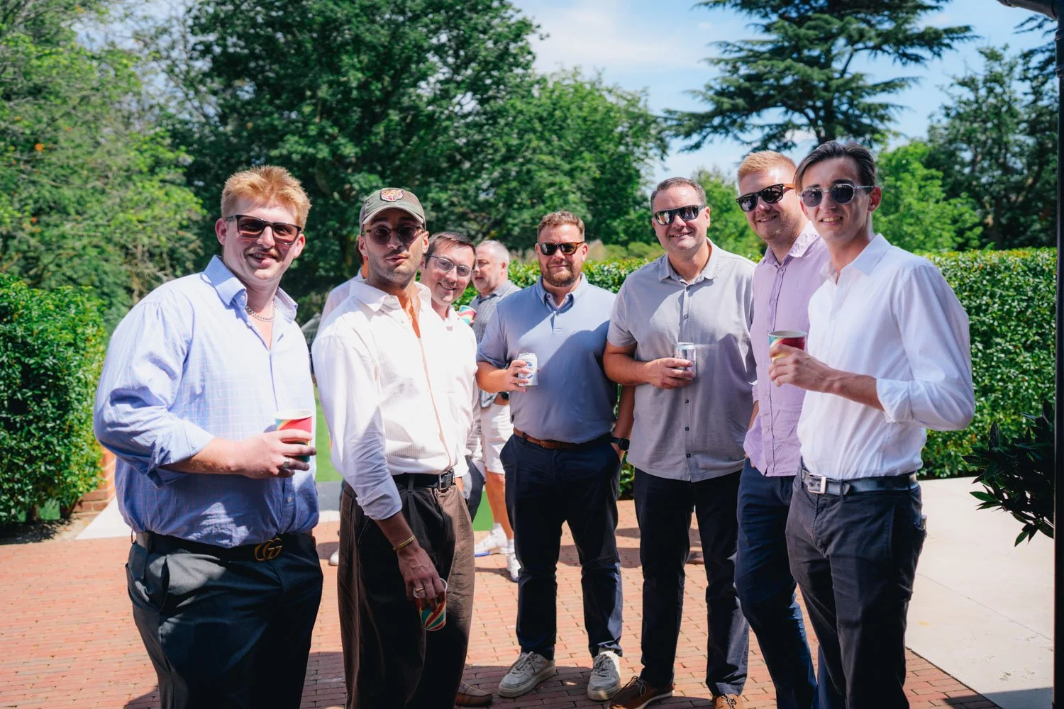 A group of eight men standing outdoors on a sunny day, smiling, wearing sunglasses and casual dress, holding drinks, with greenery and trees in the background.