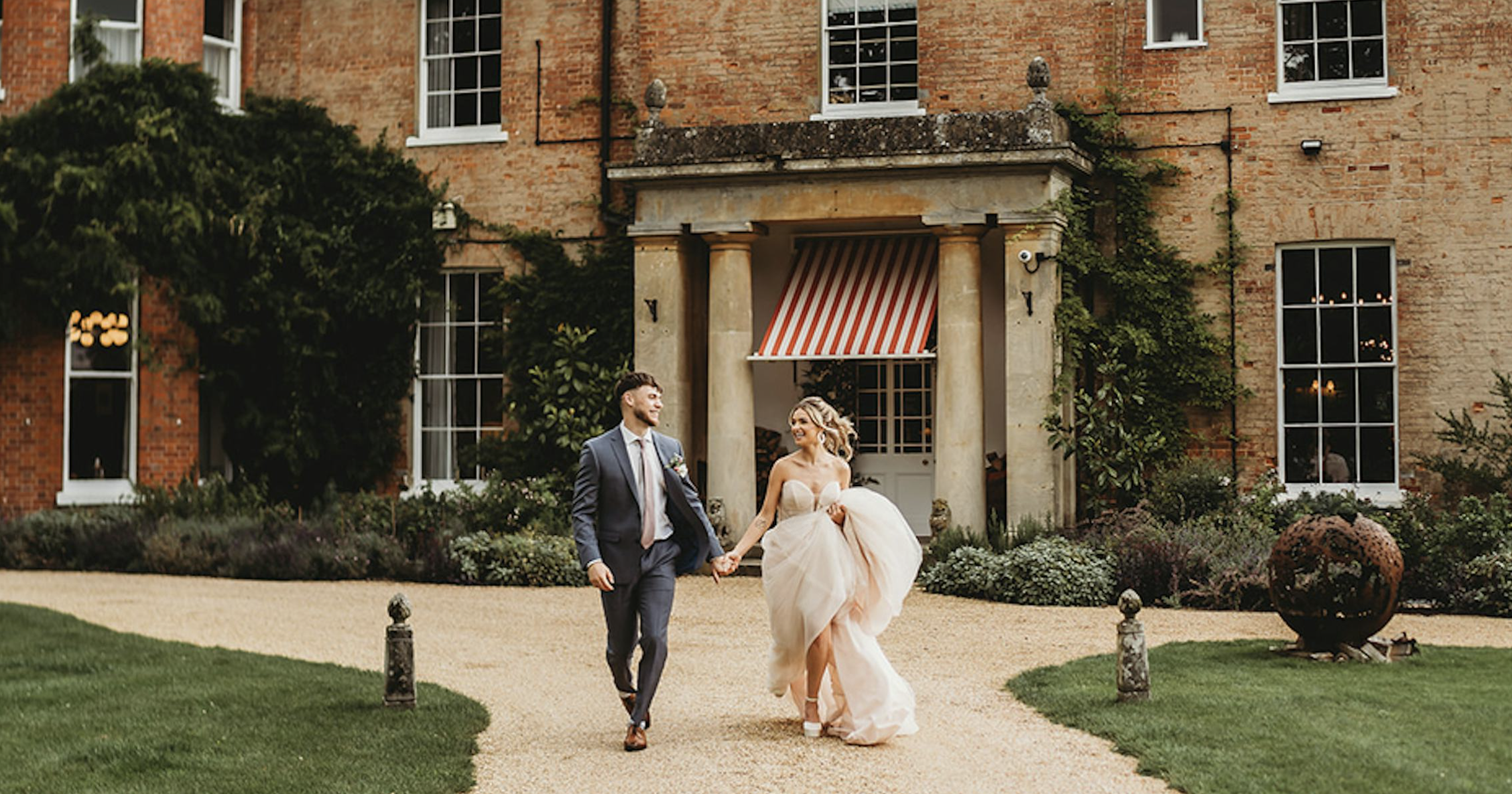 A newlywed couple walking hand in hand outside a brick building with large windows, greenery, and flowers, on a well-maintained lawn.