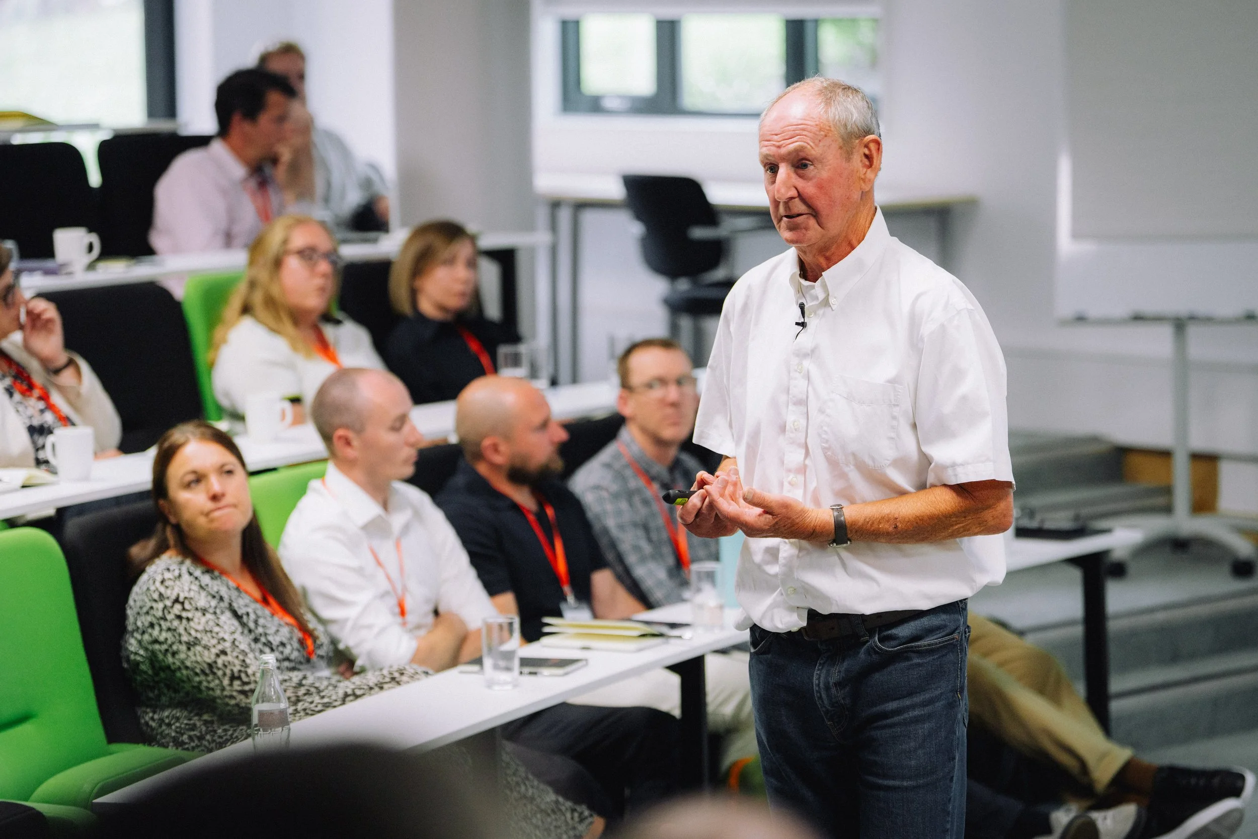 A man speaks to a seated audience in a bright lecture hall. Audience members listen attentively, some taking notes.