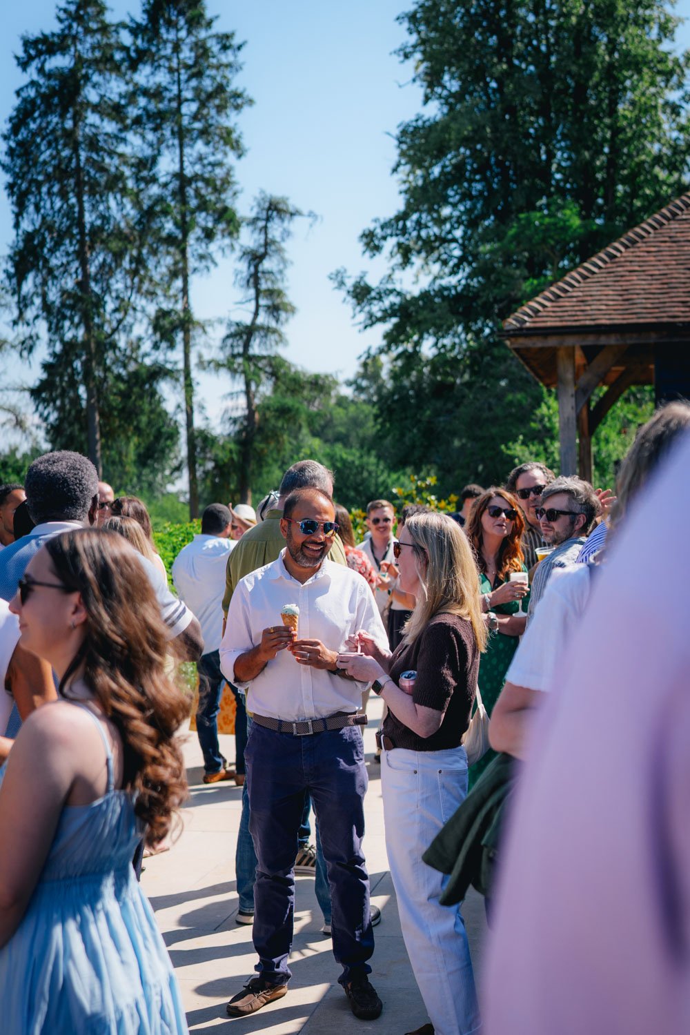 Group of diverse people enjoying a sunny outdoor gathering, some holding ice cream cones and drinks, surrounded by trees and a pavilion in the background.