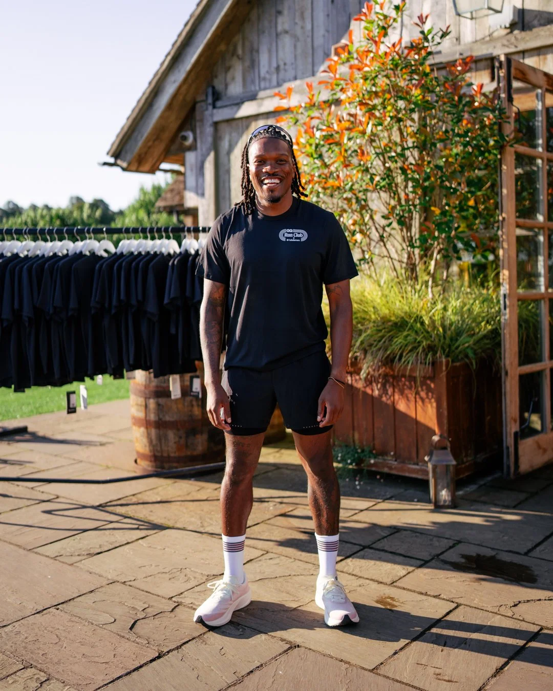 A man standing outdoors on a stone patio, smiling, wearing a black T-shirt with a logo, black shorts, white athletic shoes, and striped socks, with a barn-like building and a rack of black T-shirts in the background.