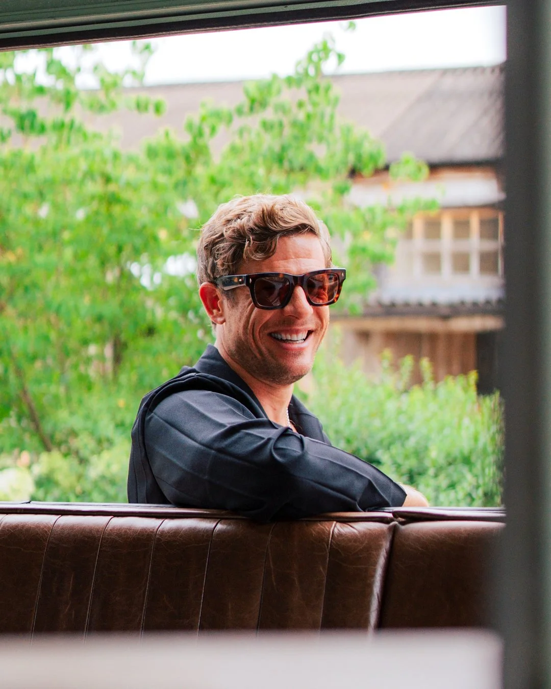 A man with curly hair and sunglasses smiling, sitting indoors near a window with greenery outside.