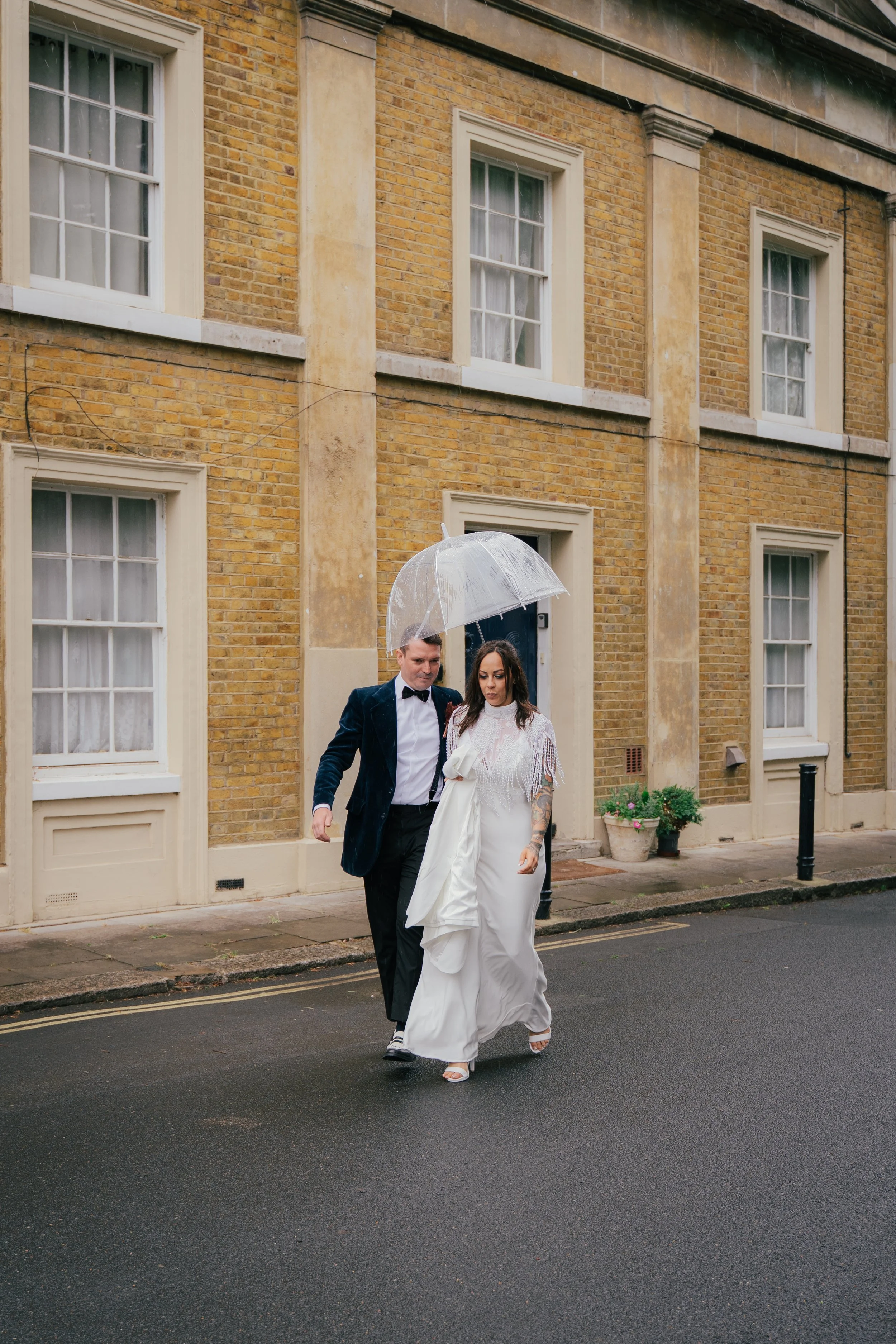 A couple dressed in wedding attire walking under a clear plastic umbrella on a rainy street in front of a yellow brick building with multiple windows.