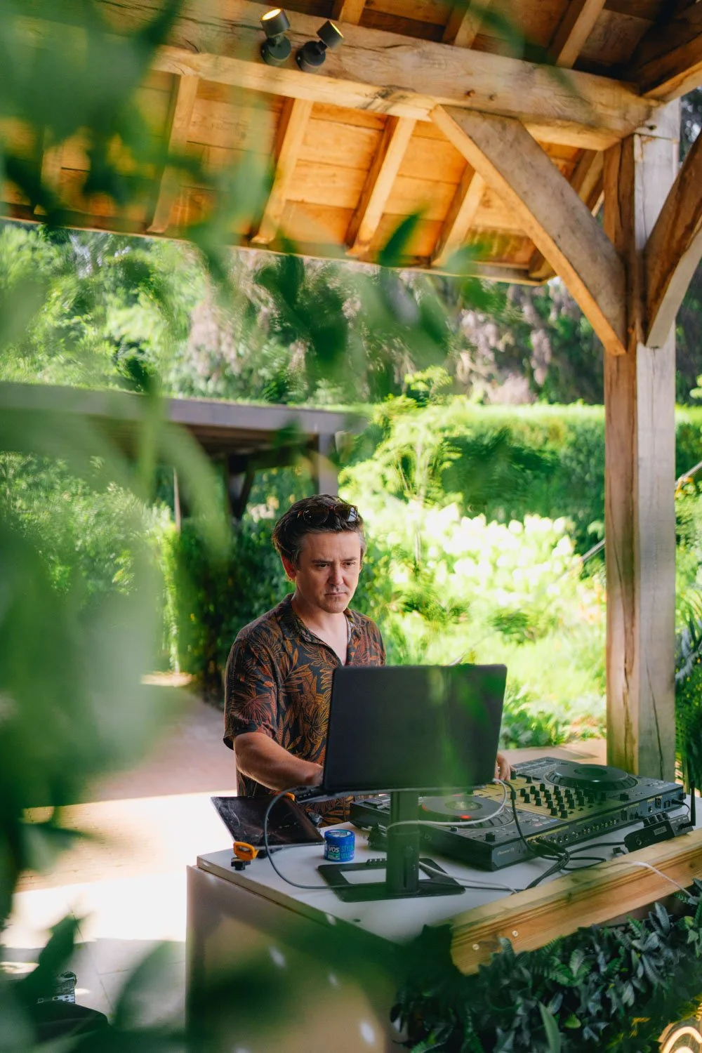 A man standing outdoors at a DJ setup under a wooden pavilion surrounded by greenery.