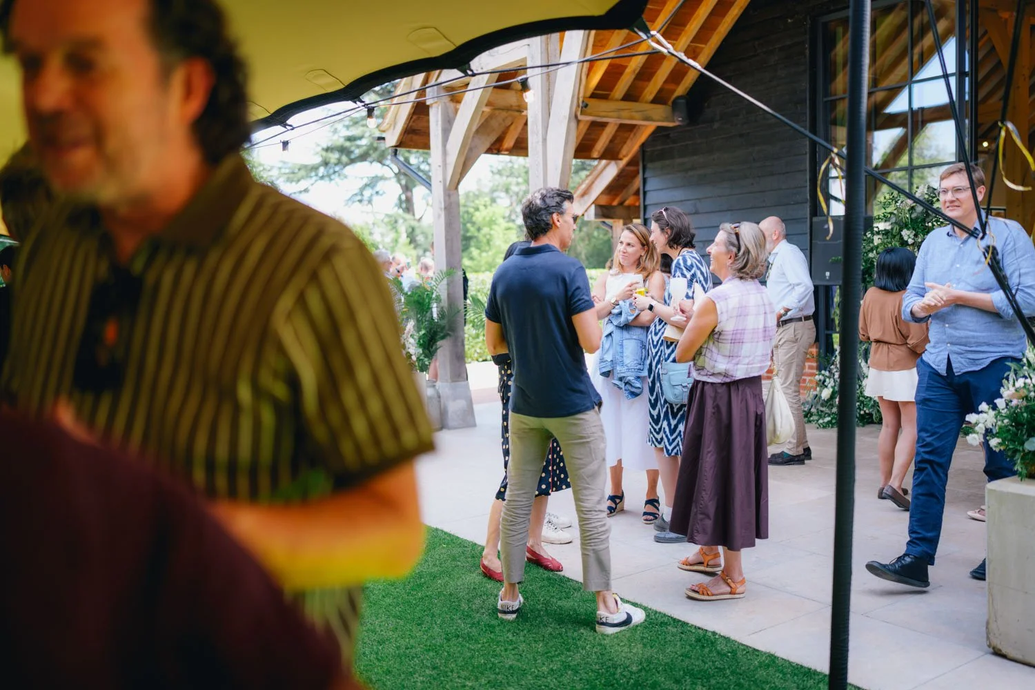 A group of people socializing outdoors on a patio with greenery and a wooden structure in the background, with one person close to the camera partially out of focus.