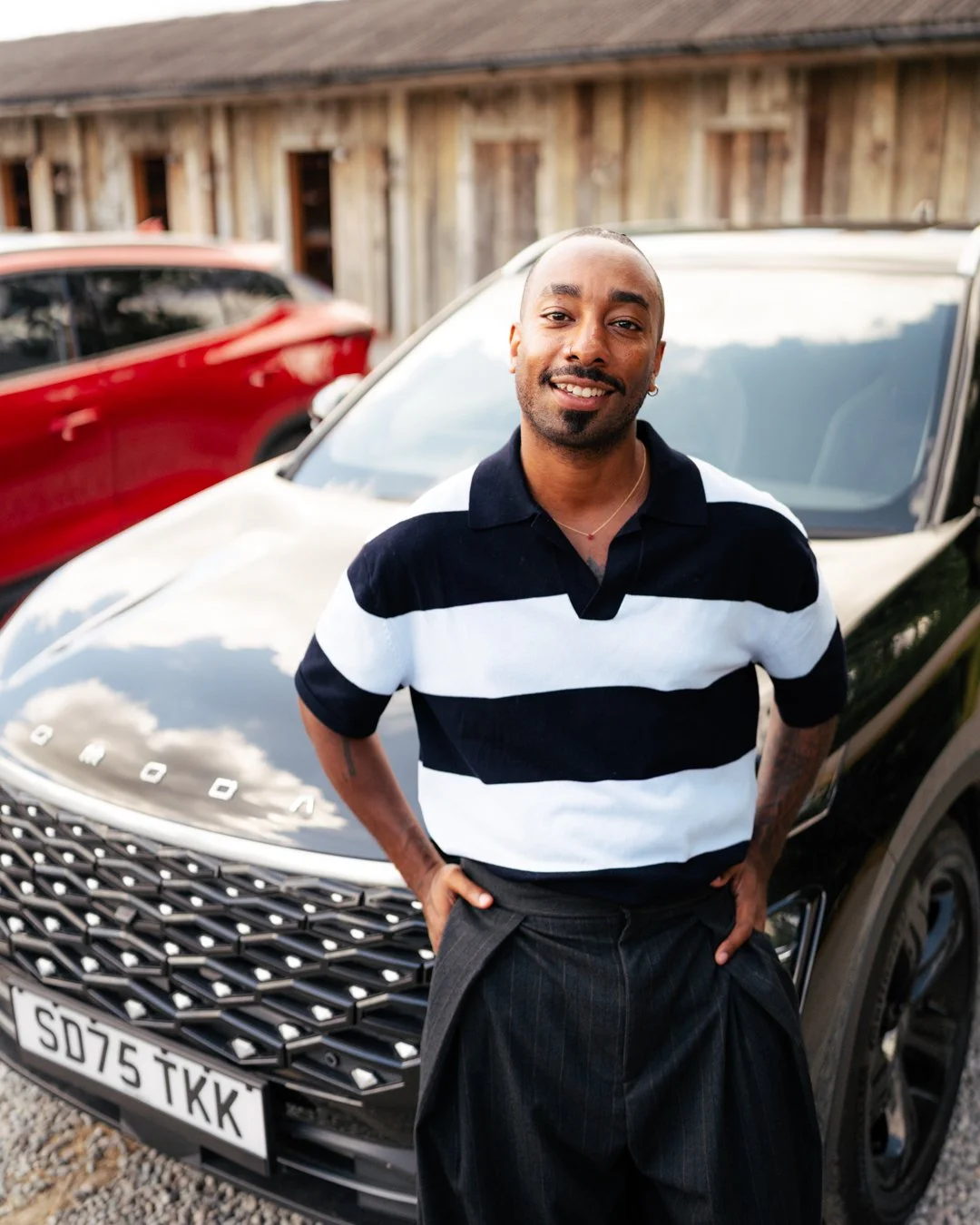A man standing outdoors in front of a black Range Rover, wearing a black and white striped polo shirt and black pants, smiling at the camera.