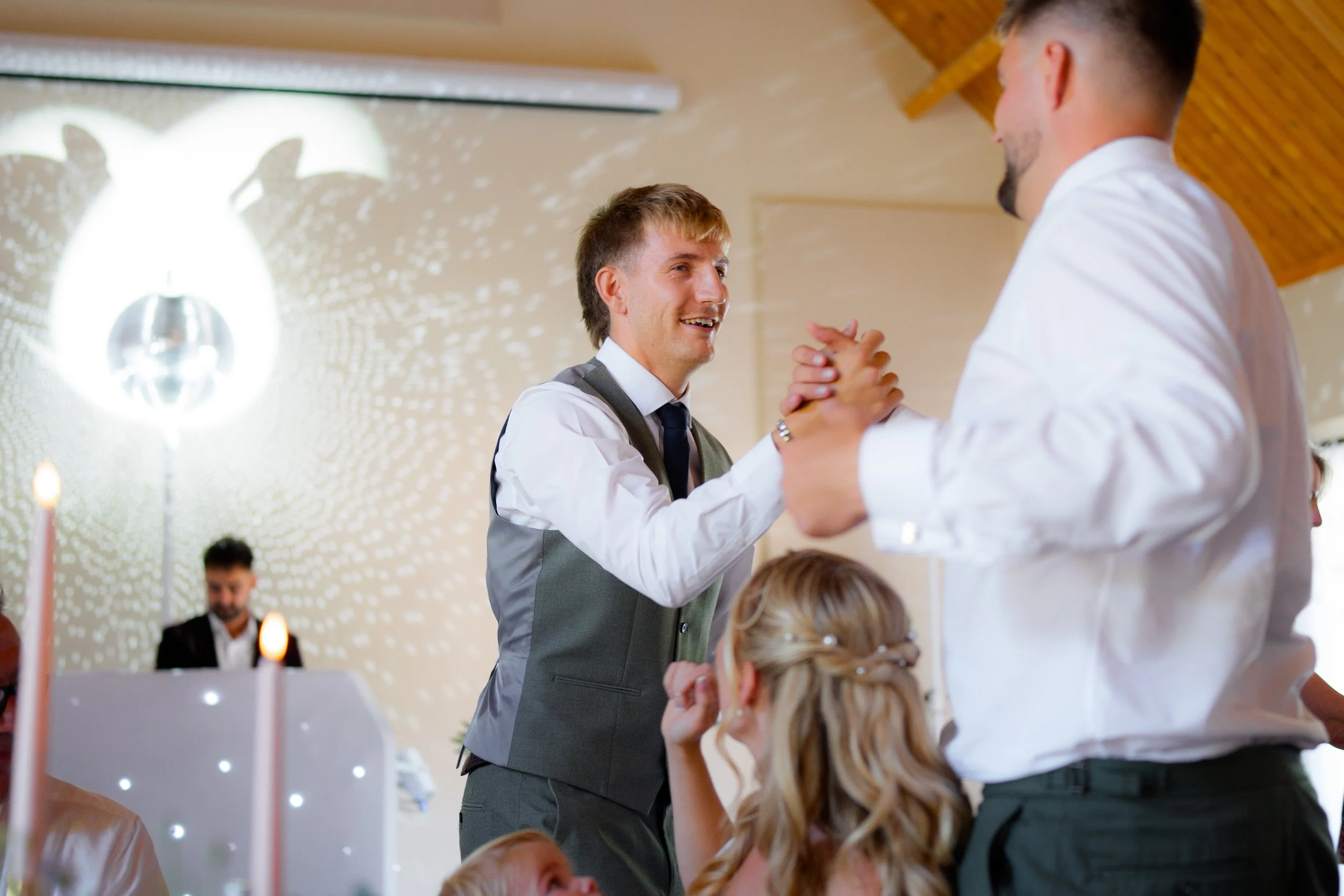 A man with a beard and tie, wearing a suit, is shaking hands with another man in a white shirt at a wedding reception. Several children are in the foreground, and a woman with blonde hair is sitting nearby. The background has wedding decorations and 