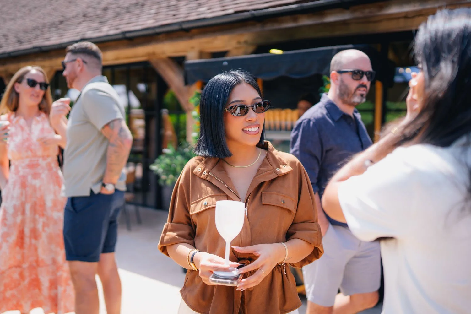 Group of people socializing outdoors, woman in center holding a wine glass, wearing sunglasses and a brown jacket, smiling and talking to another woman.