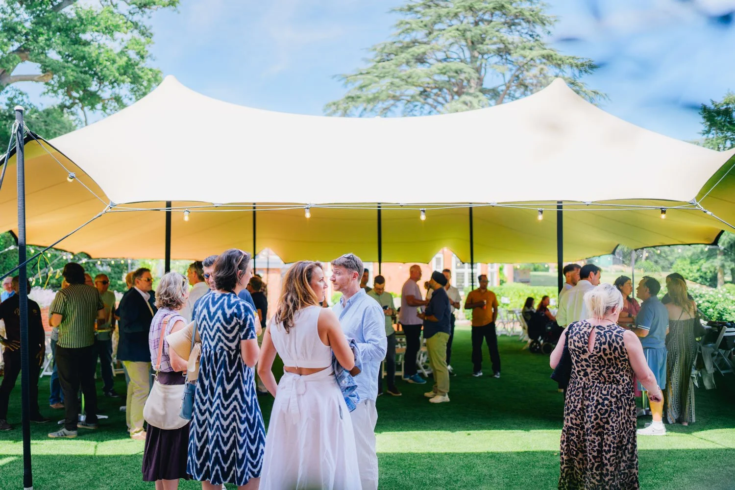 People gathering and socializing under a large outdoor canopy tent in a park on a sunny day.
