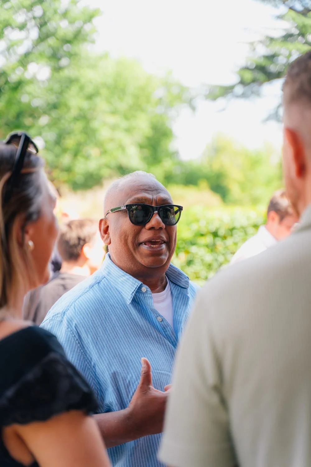 Man with sunglasses wearing a light blue striped shirt talking to a group of people outdoors.