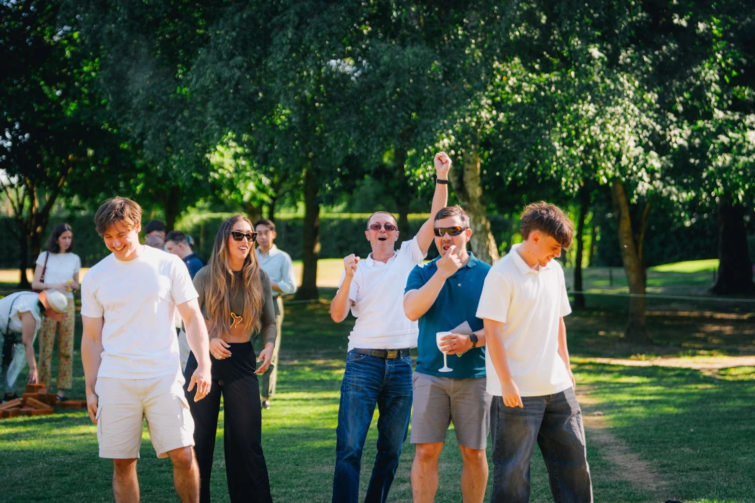 Group of young adults enjoying an outdoor celebration in a park, some dancing and laughing, with trees and sunlight in the background.