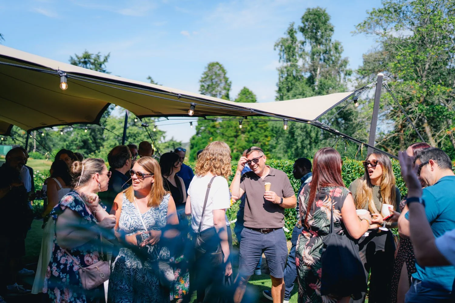 Group of people at an outdoor party under a canopy, socializing and enjoying ice cream on a sunny day surrounded by greenery.