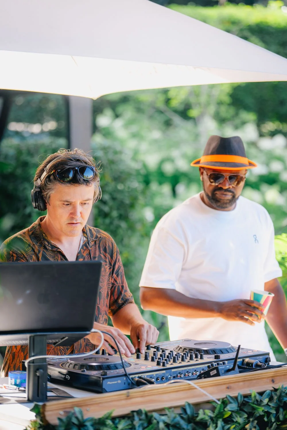 Two men at an outdoor DJ setup surrounded by greenery. One man, with headphones and sunglasses on his head, is operating a DJ controller. The other man, wearing a hat with an orange band and sunglasses, is holding a cup and watching.