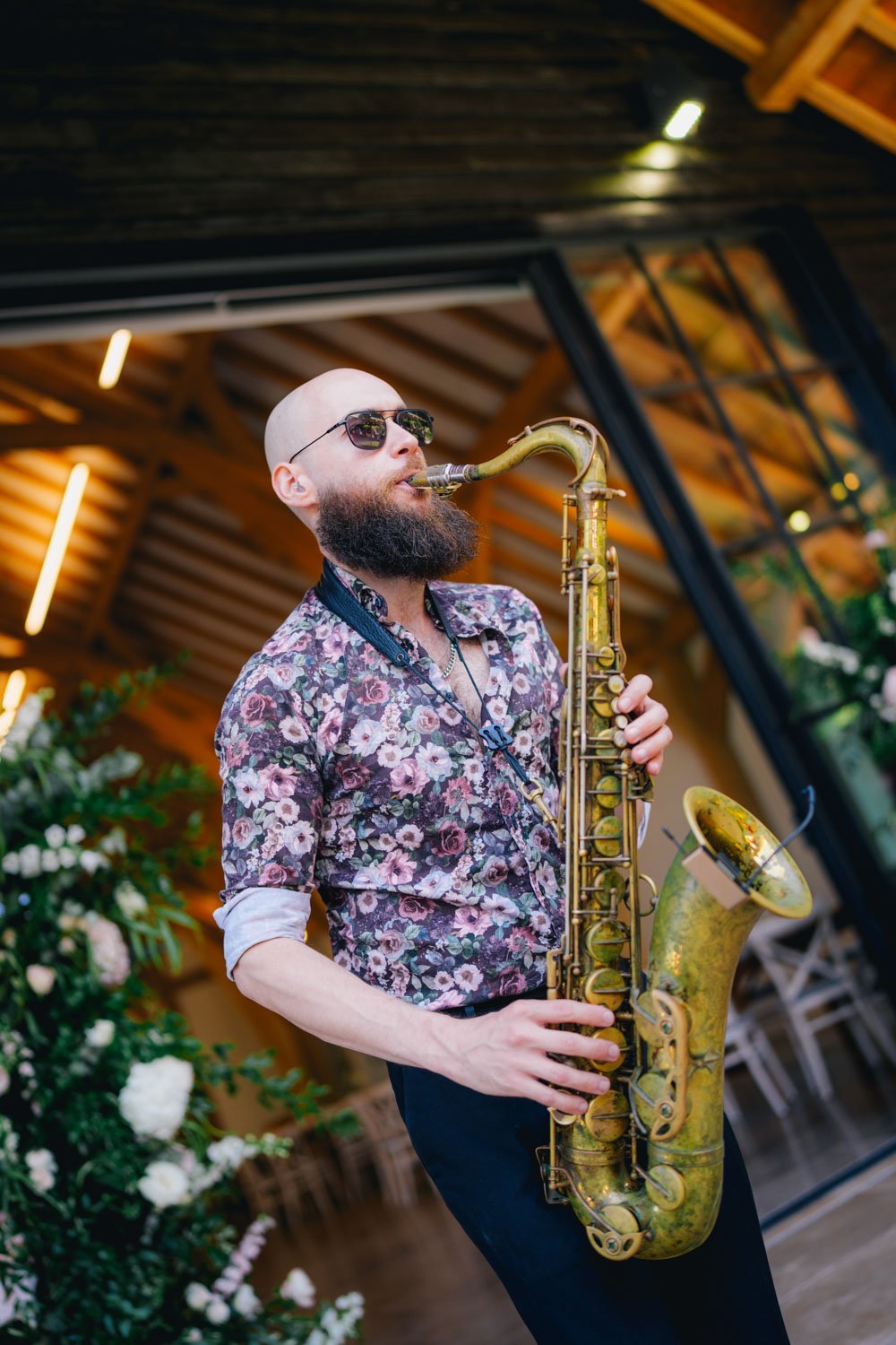 A man with a bald head, beard, sunglasses, and a floral shirt playing a saxophone indoors with wooden ceiling and plants in the background.