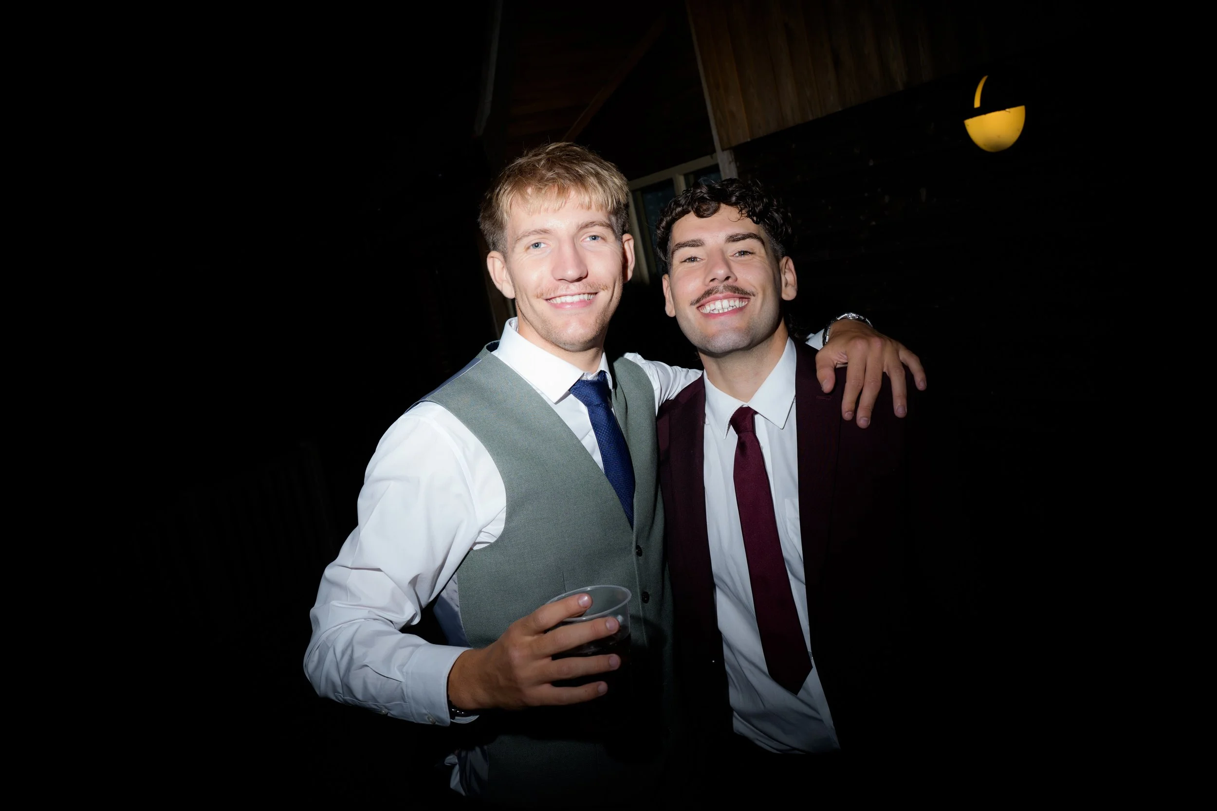 Two men dressed in suits smiling and standing close, one with their arms around the other's shoulder, at an indoor event.