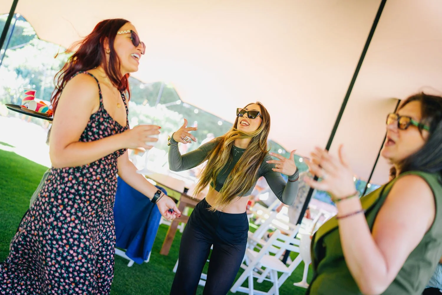 Three women dancing and laughing under a tent at an outdoor party.