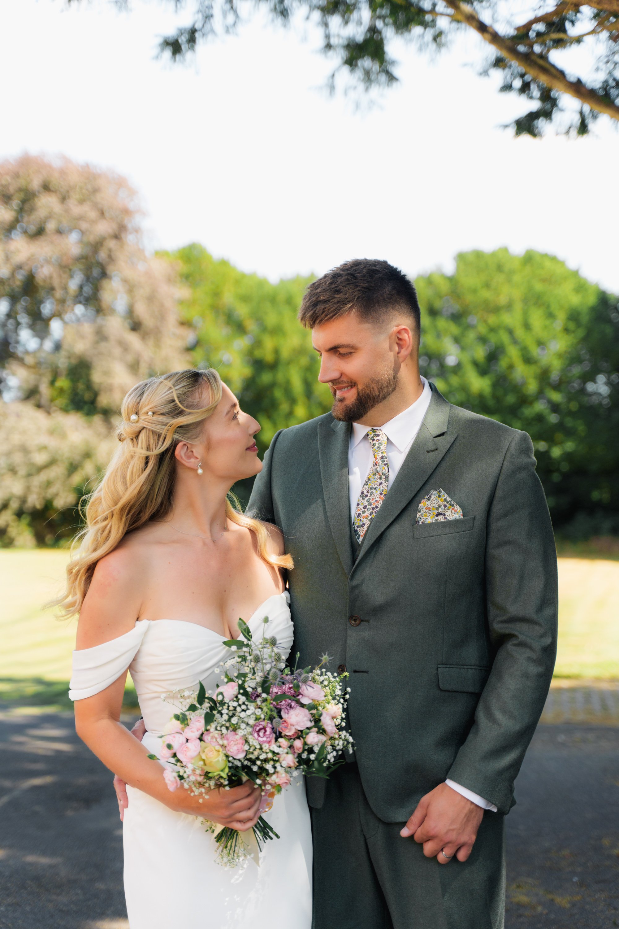 A bride and groom standing close together outdoors, with the bride holding a bouquet of pink and white flowers and the groom wearing a dark gray suit with a patterned tie, surrounded by trees and greenery.