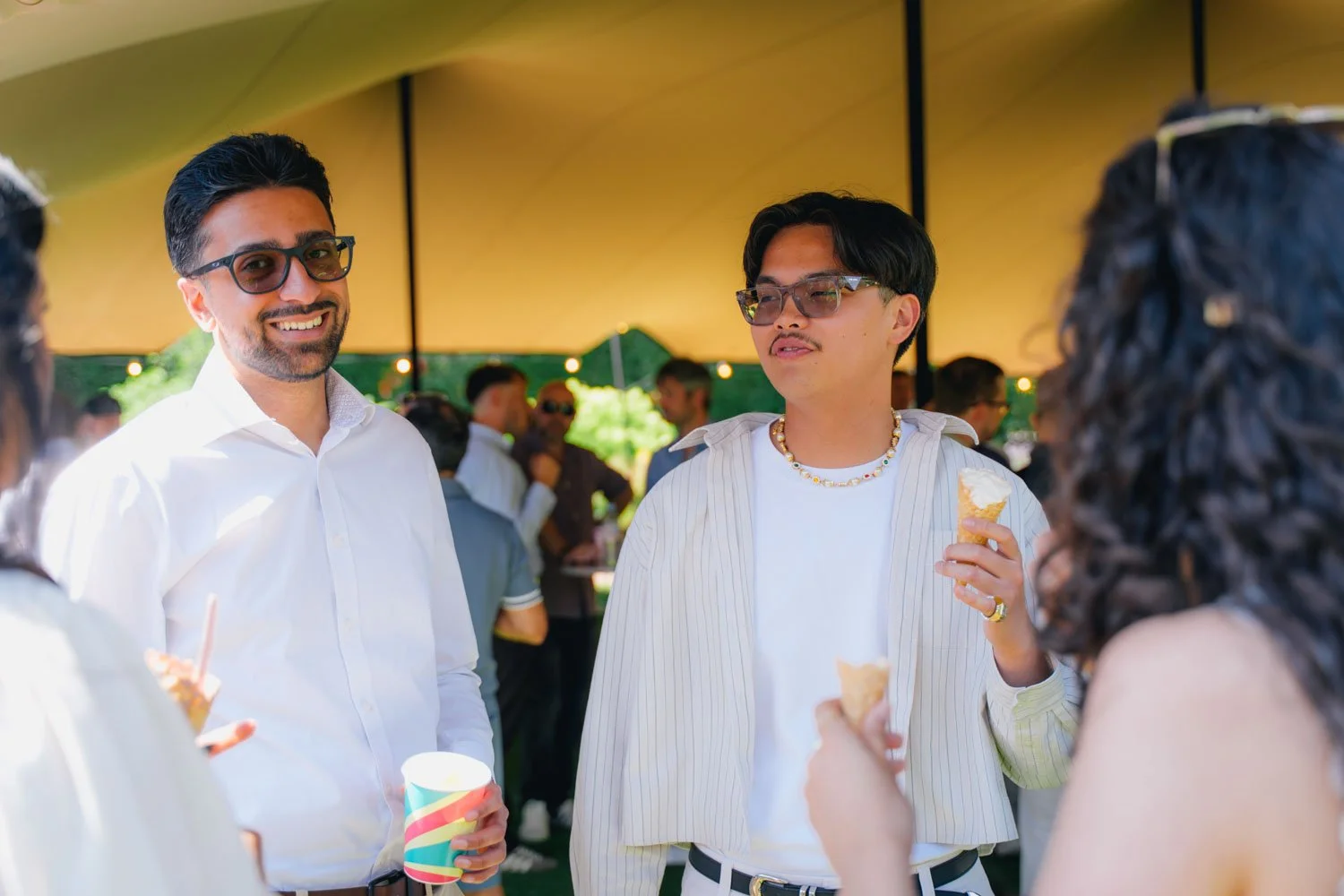 People at an outdoor party or gathering, socializing under a canopy, holding snacks and drinks.