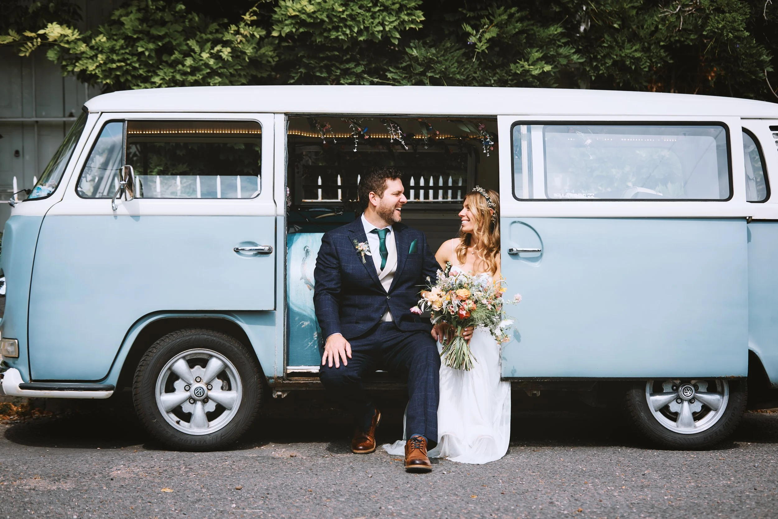 A bride and groom sitting on the bumper of a vintage blue Volkswagen van, smiling at each other. The bride holds a bouquet of flowers, and they are outdoors with greenery in the background.