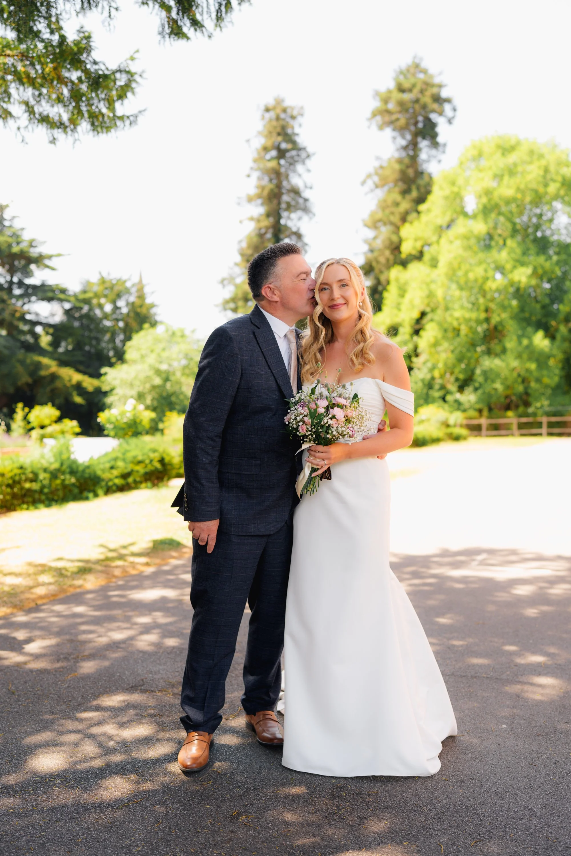 A newlywed couple outdoors with tall trees and green foliage in the background. The groom, wearing a dark checkered suit and brown shoes, kisses the bride on her forehead. The bride, in a white off-shoulder wedding dress, holds a bouquet of pink and 