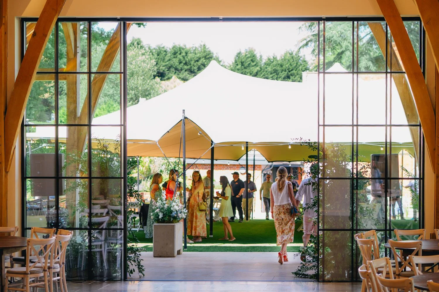 People gathered outdoors at a garden event seen through large glass doors from inside a building.
