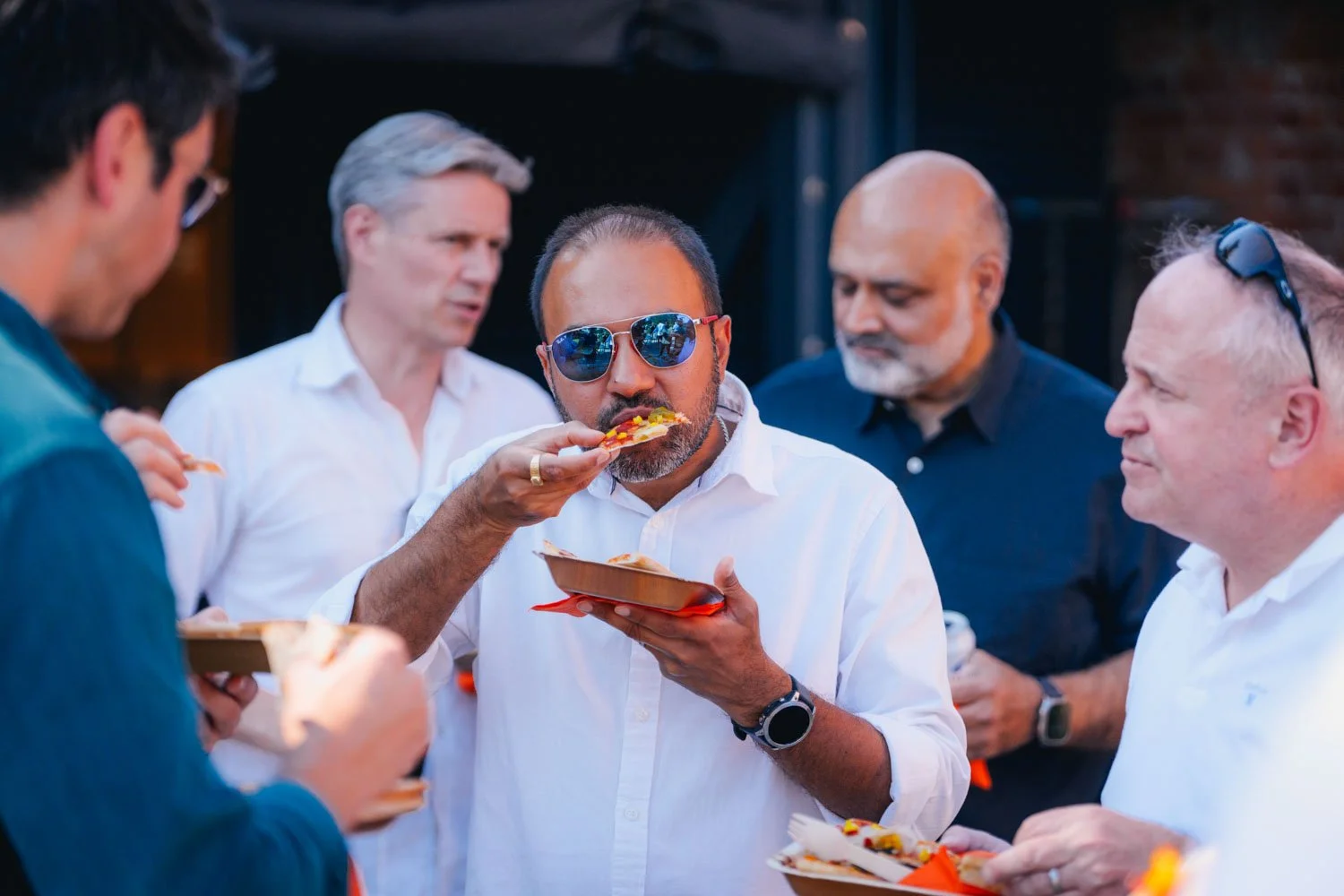 Group of men gathered outdoors, one wearing sunglasses and eating pizza, others holding plates with food.