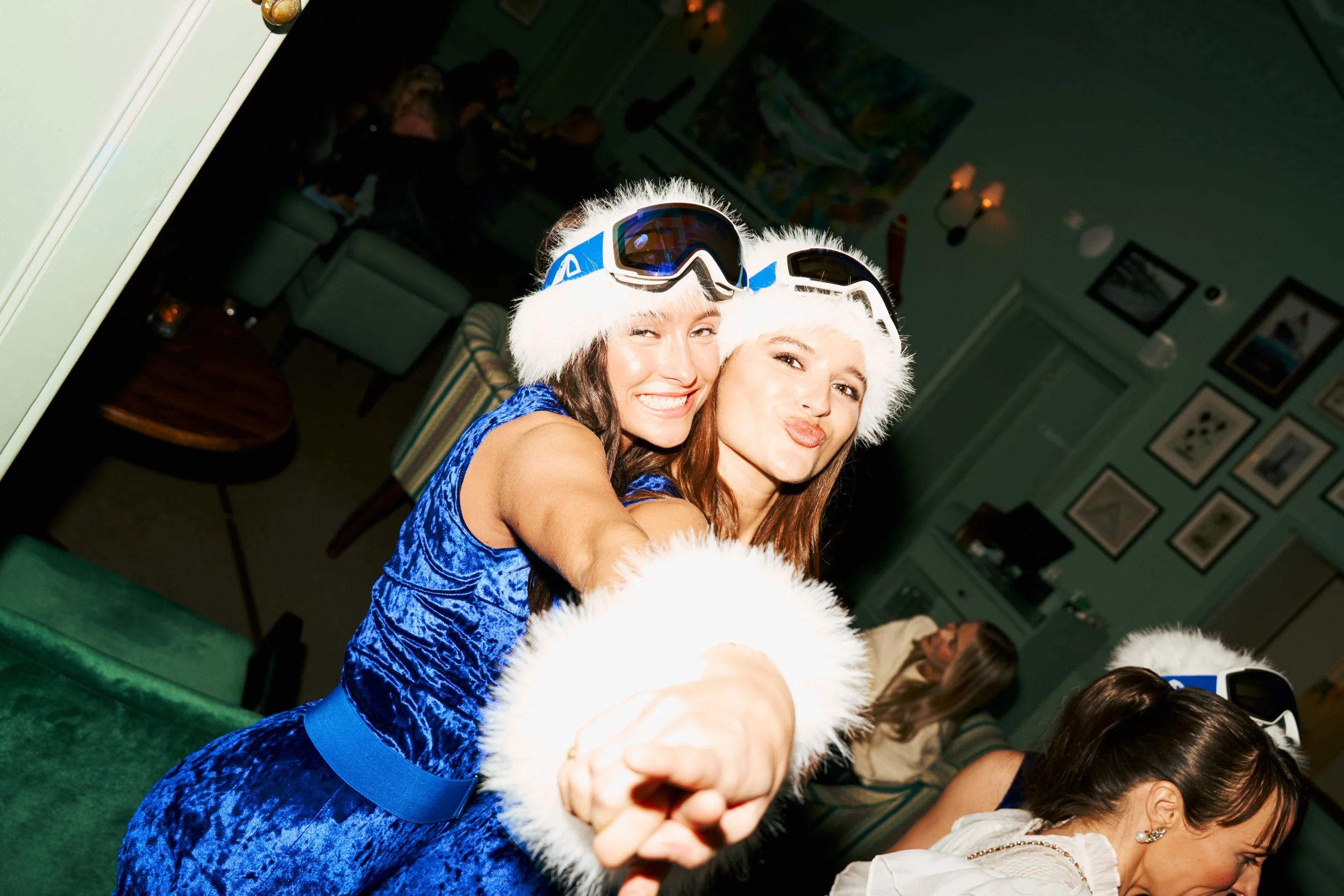 Two women wearing blue velvet dresses and white faux fur Santa hats with ski goggles on their heads, smiling and posing for a photo at a holiday party.