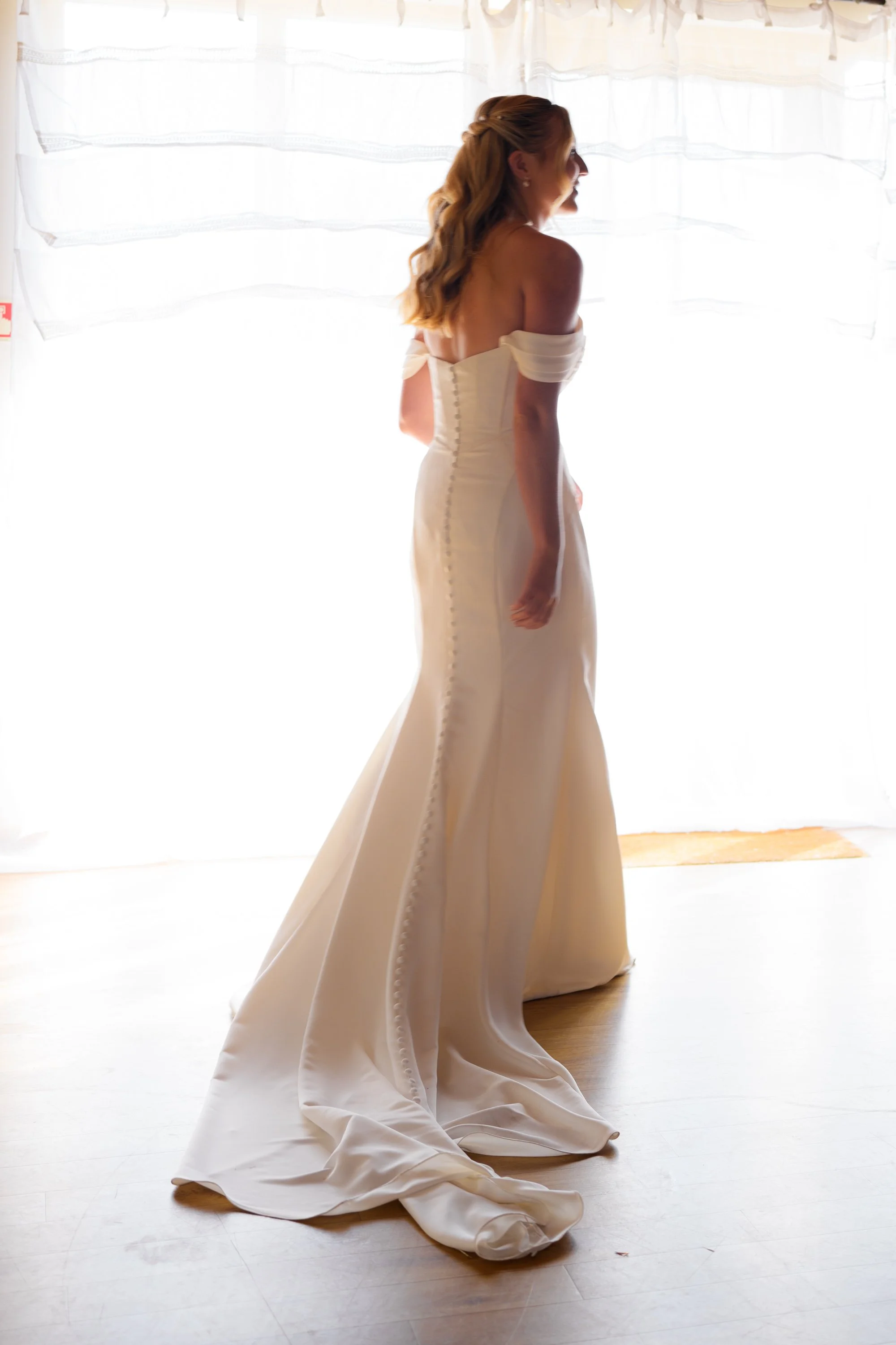 A woman wearing a white, strapless wedding dress. Her long, wavy hair is styled back. She is standing indoors on a wooden floor in front of sheer white curtains that let in natural light, with her head turned to the side, showing her profile.
