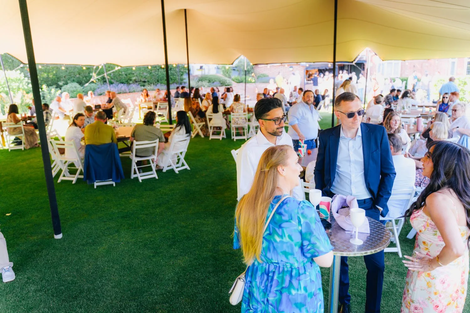 People gathered under a large yellow canopy at an outdoor event, with some standing and socializing in the foreground, and others seated at tables in the background on a grassy area.