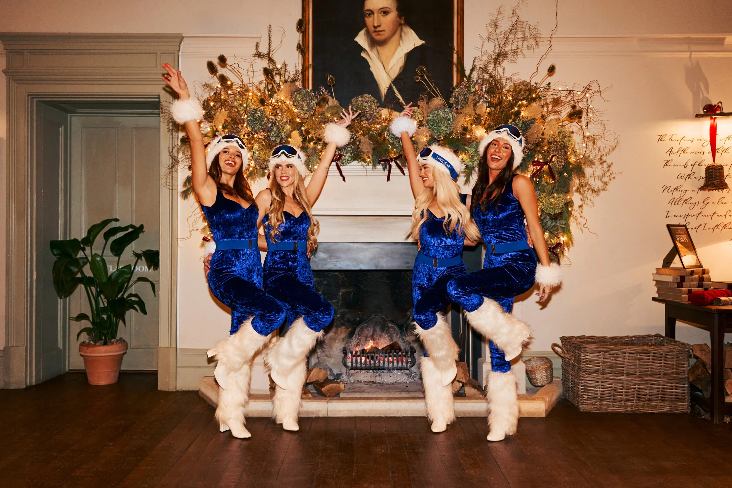 Four women dressed in matching blue velvet outfits with white faux fur trim and boots, standing in front of a fireplace decorated with flowers and lights, with a portrait hanging above. They are smiling, with two raising their arms, celebrating.