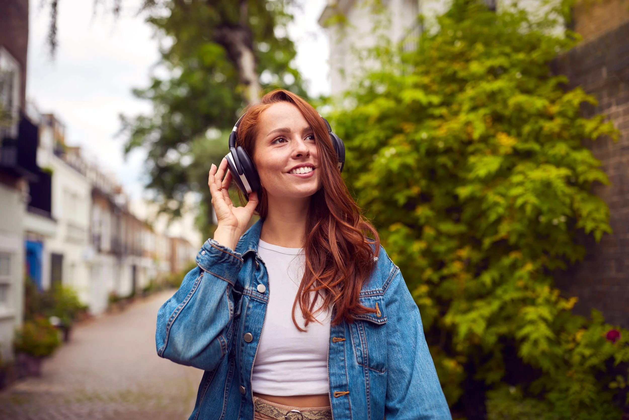 A young woman with long red hair wearing a denim jacket and white crop top, smiling and listening to music with large black headphones outdoors in a leafy neighborhood.