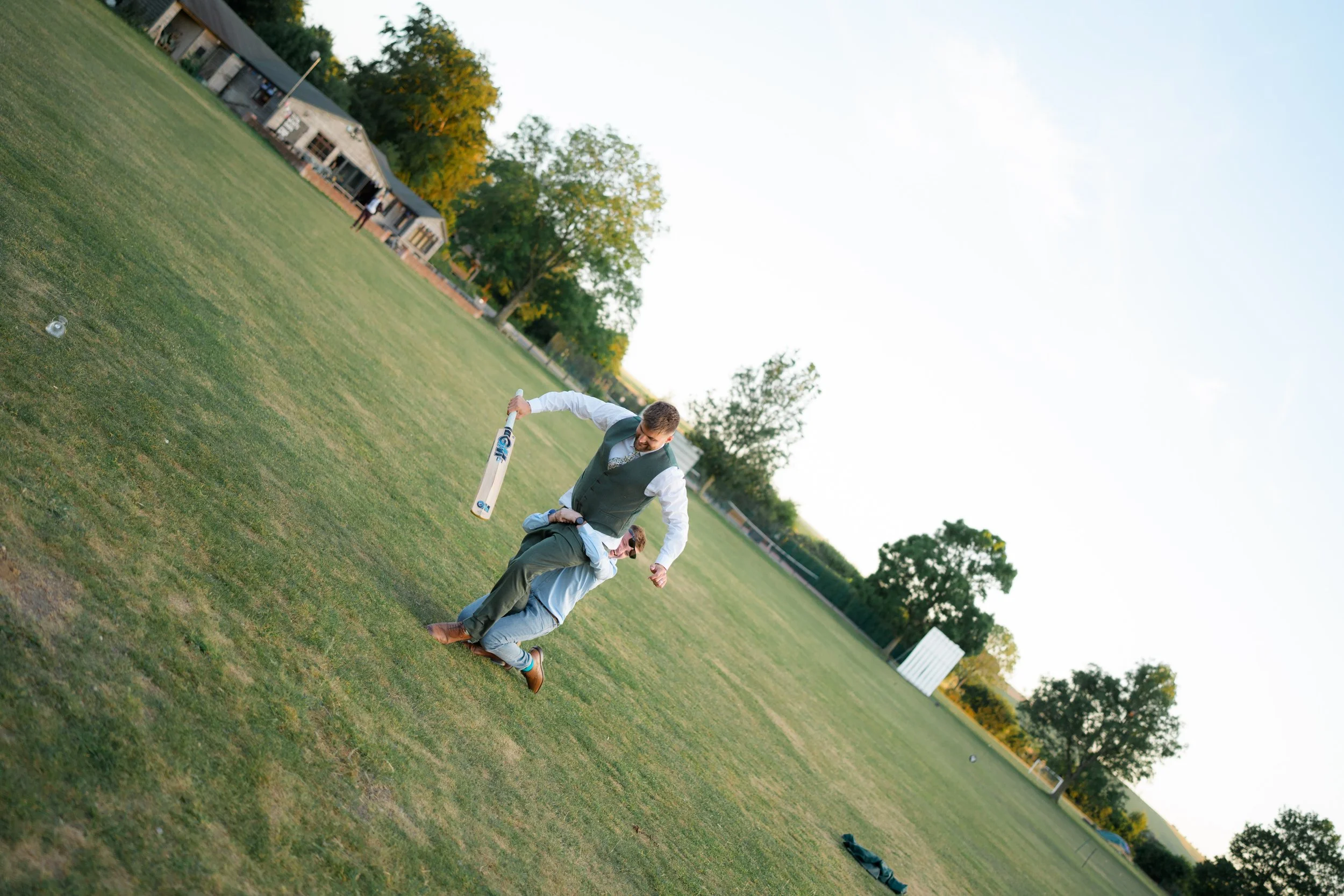 Two men are in a park, one is bending down and the other is standing, holding a cricket bat, near a grassy field with trees and some buildings in the background during what appears to be late afternoon or early evening.