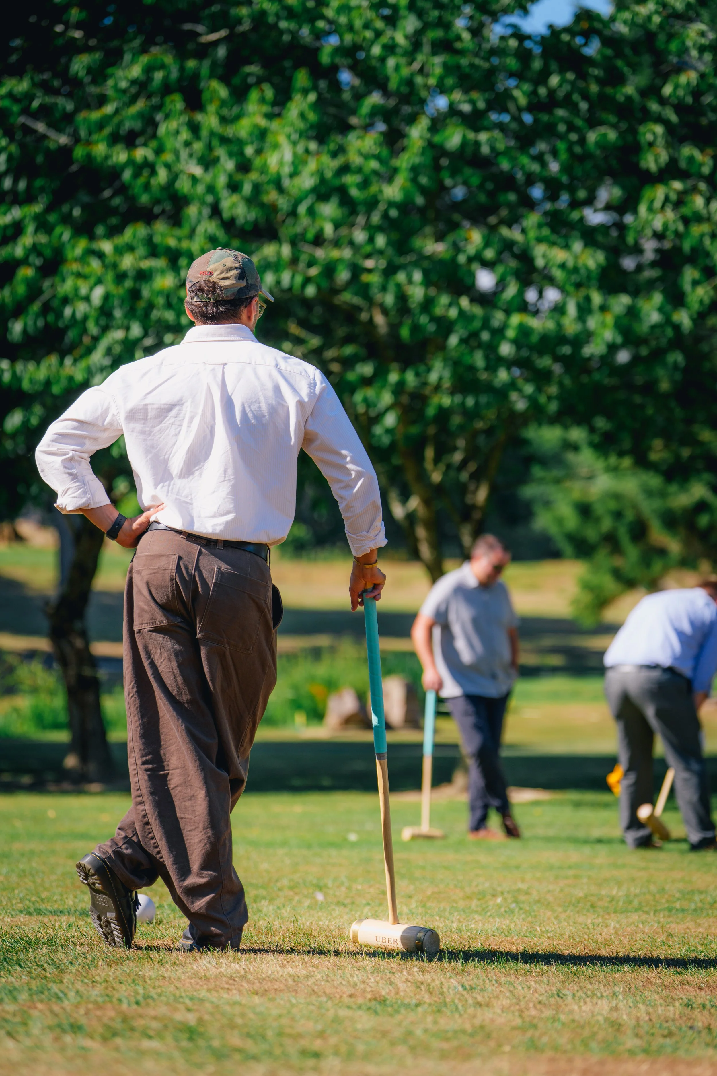 A person in a white shirt holds a croquet mallet on a grassy lawn, with others playing in the background.
