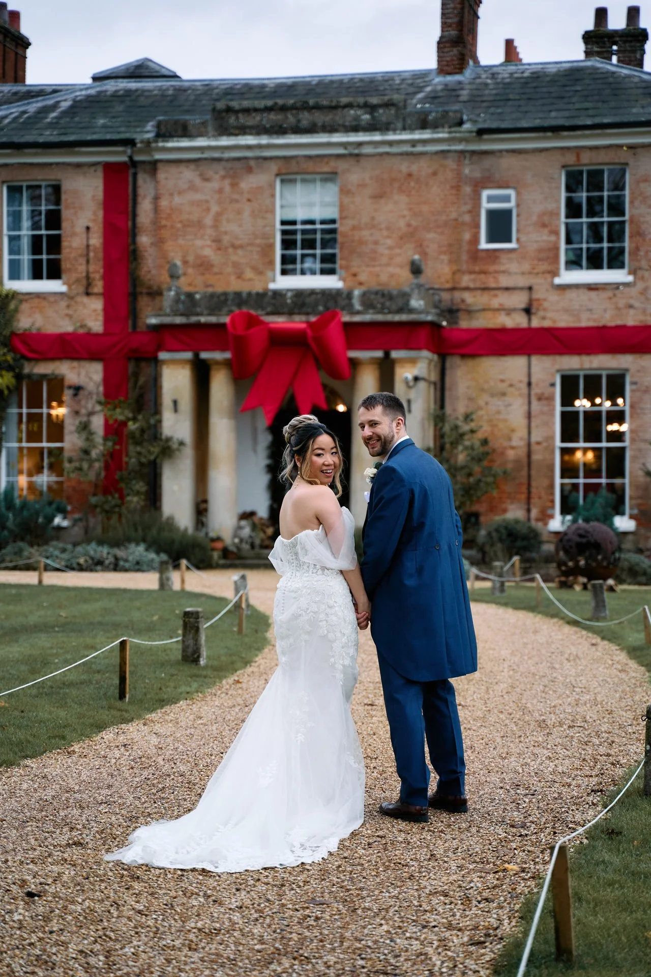A bride and groom holding hands and looking at the camera outside a red brick building decorated with a large red bow and ribbon for a wedding celebration.