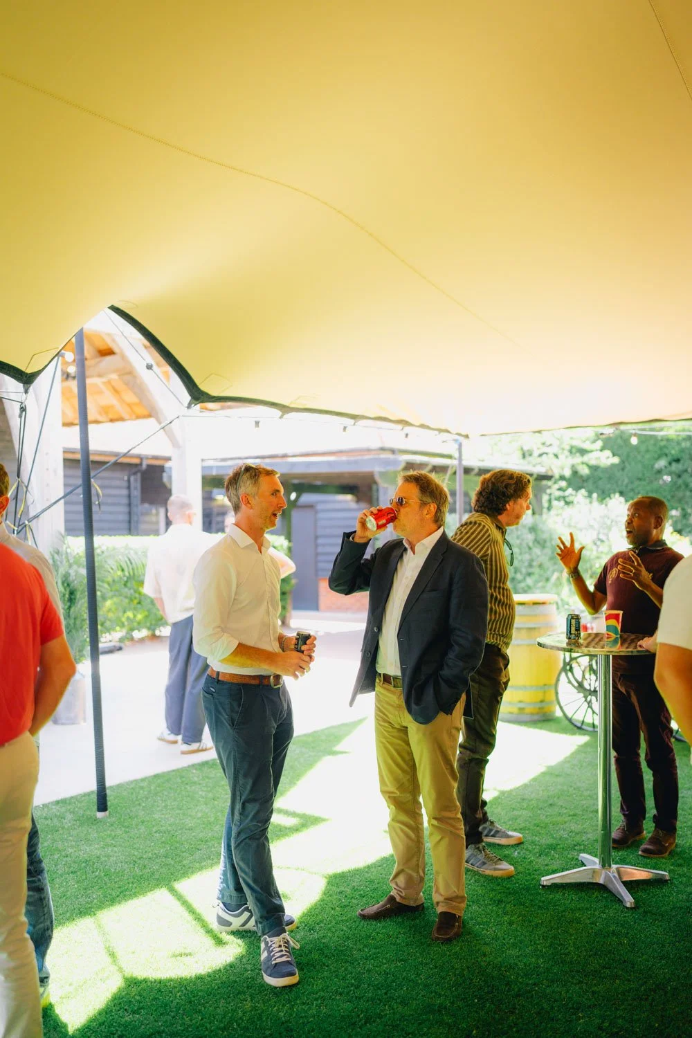 People socializing at an outdoor gathering under a yellow canopy, with some holding drinks and engaging in conversation.